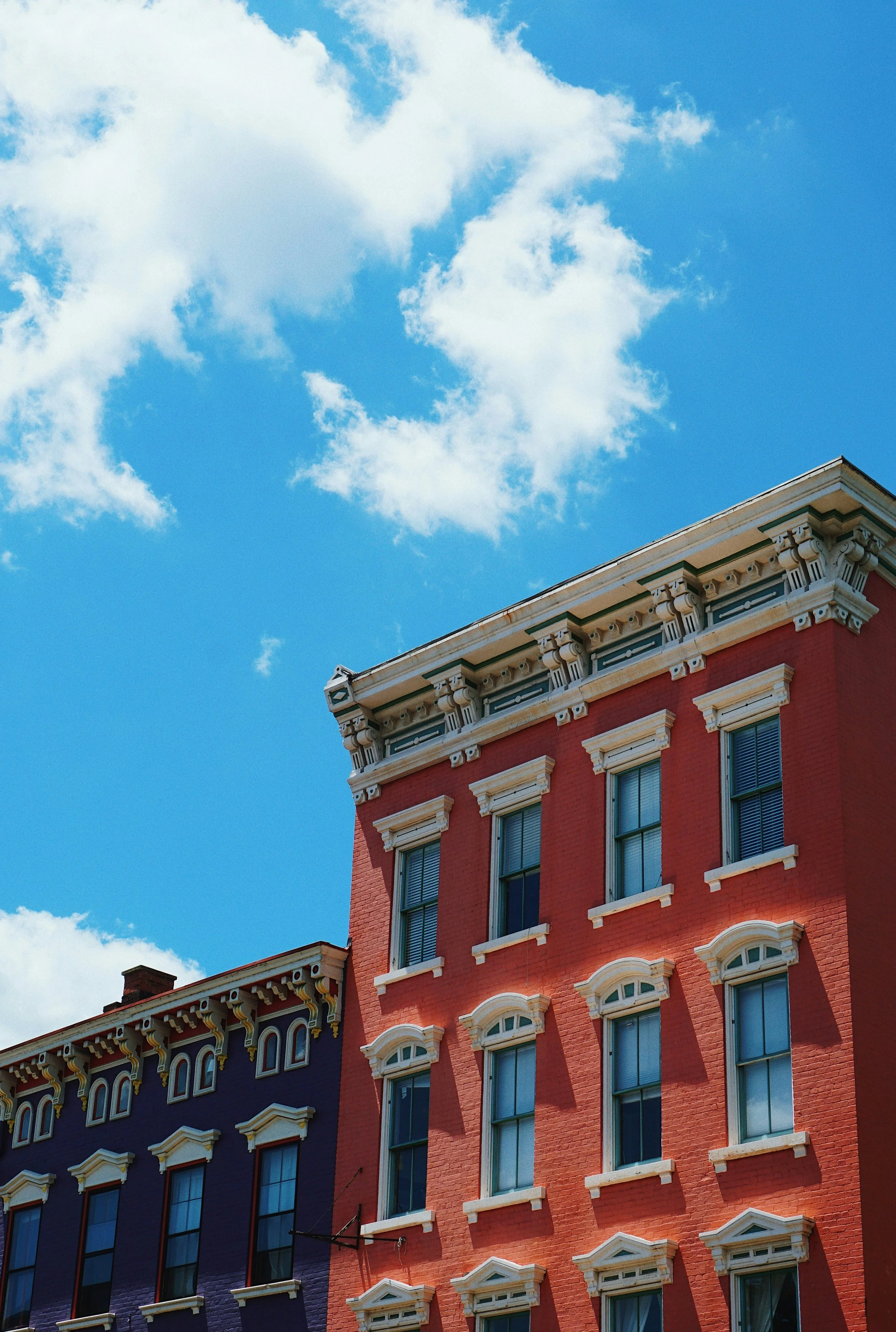 The image shows two colorful buildings with ornate architectural details, one painted purple and the other pink, with blue sky and white clouds overhead.