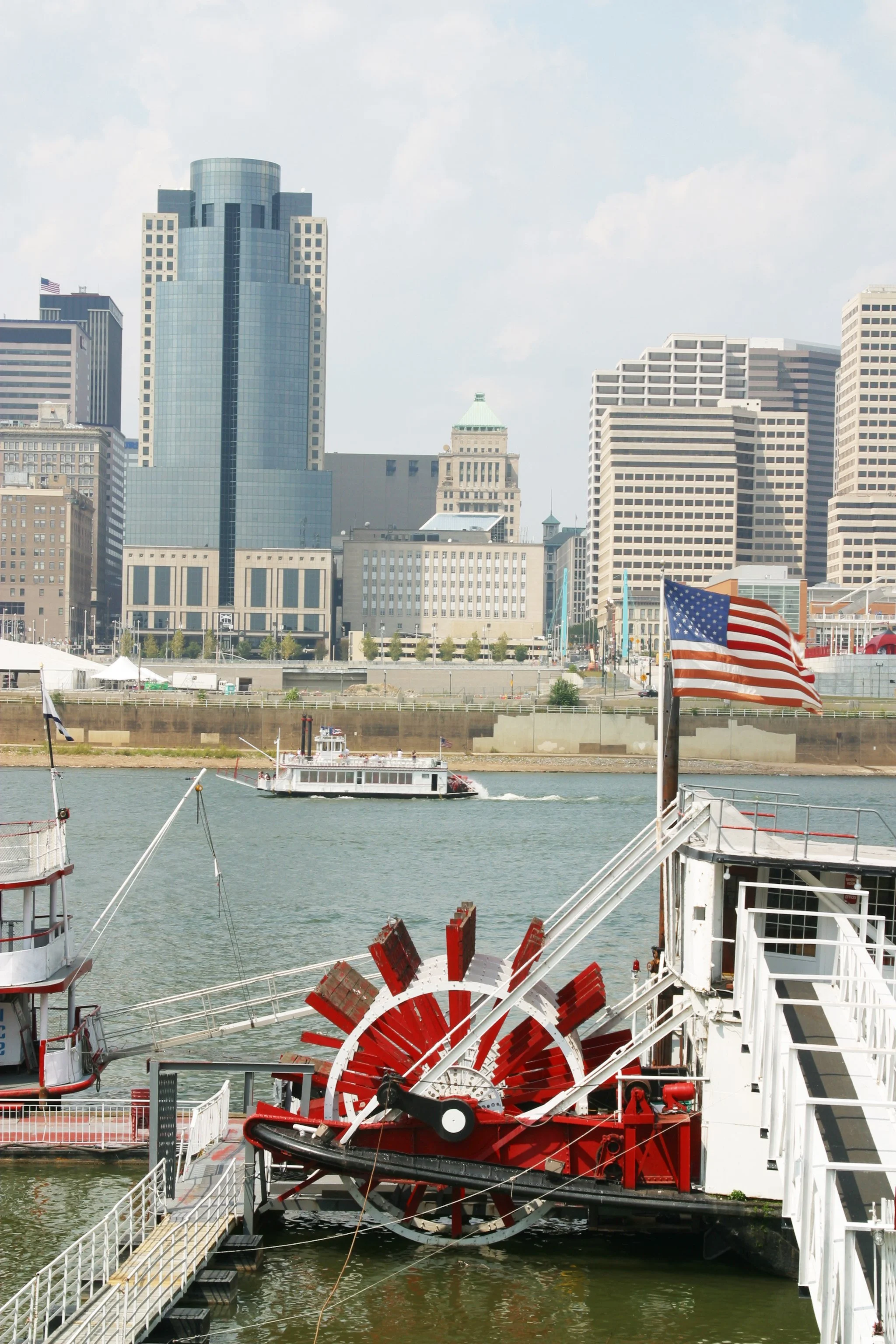 The Cincinnati city skyline with tall buildings, including a modern glass skyscraper, and the Ohio River in the foreground with boats, including a ferry boat with an American flag and a red water pump, during daytime.