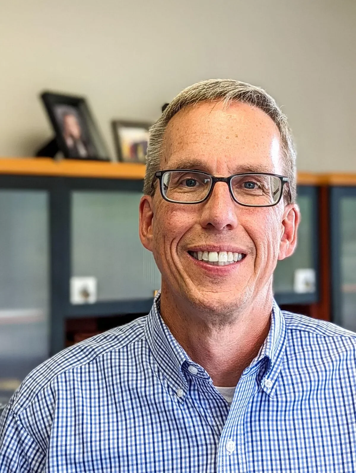 A smiling middle-aged man with short gray hair and black glasses, wearing a blue and white checkered shirt, indoors with framed photos on a bookshelf in the background.