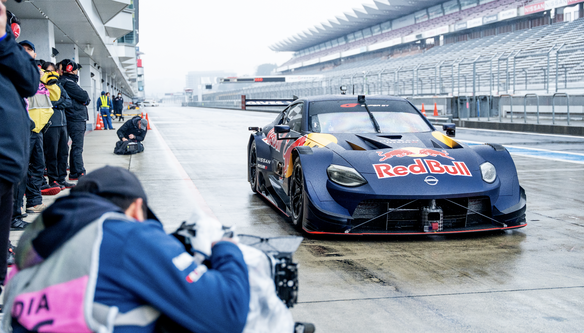 Race car in garage with team members and photographers around during a motorsport event at a racetrack.