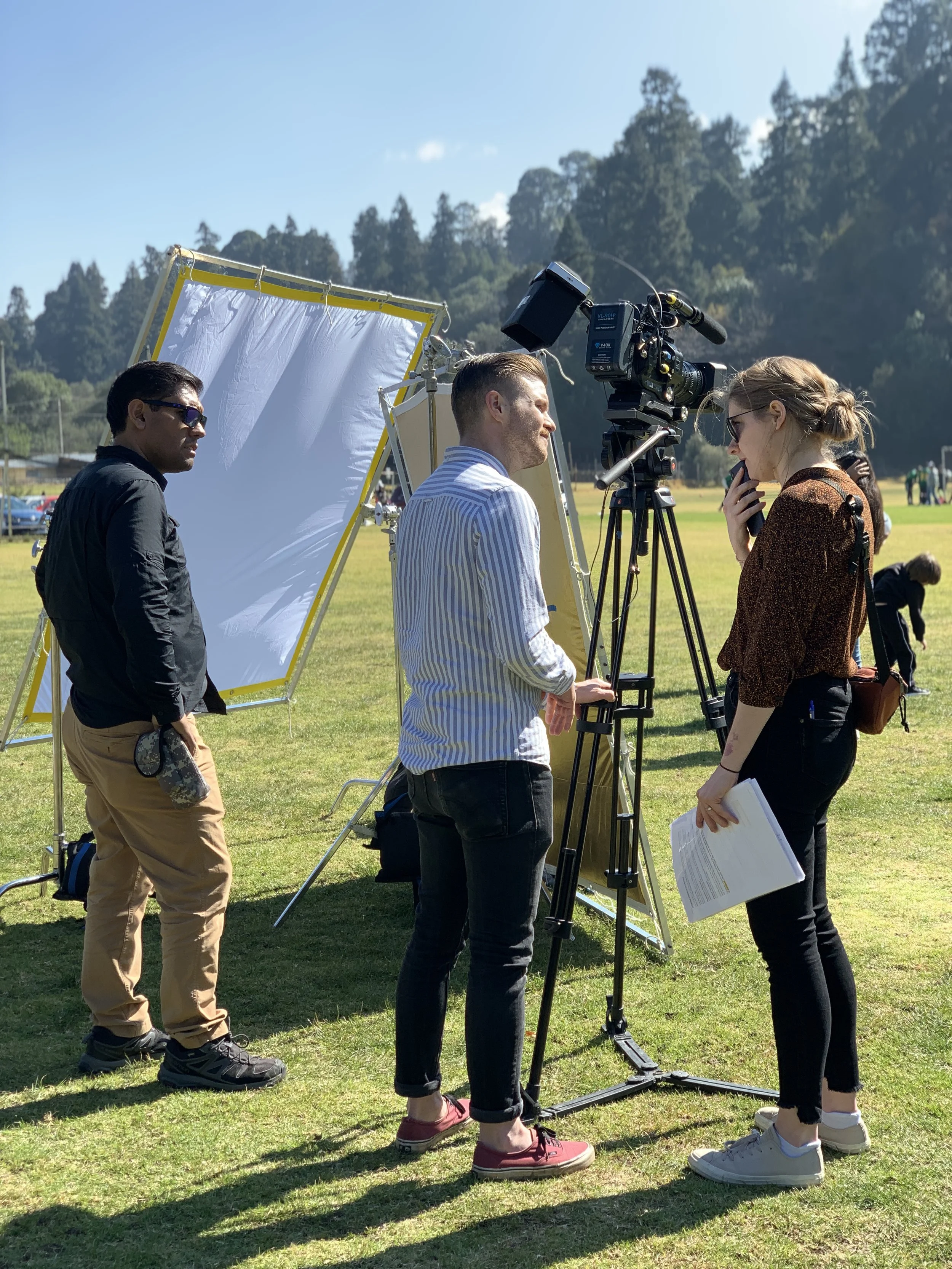 Filming crew setting up equipment outdoors with a camera on a tripod, a woman holding papers, and a woman standing near the camera, with a backdrop of trees and a grassy field.