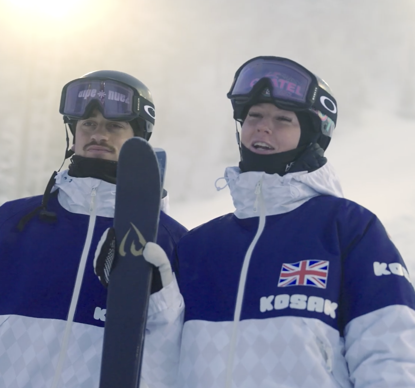 Two athletes in winter sports gear, wearing protective helmets and goggles, standing outdoors in a snowy environment. One is holding a pair of skis, and both have the British flag on their jackets.