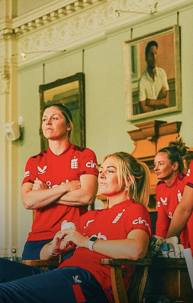 Group of female athletes in red sports jerseys, one standing with arms crossed, two seated, all inside a room with framed paintings on the wall.