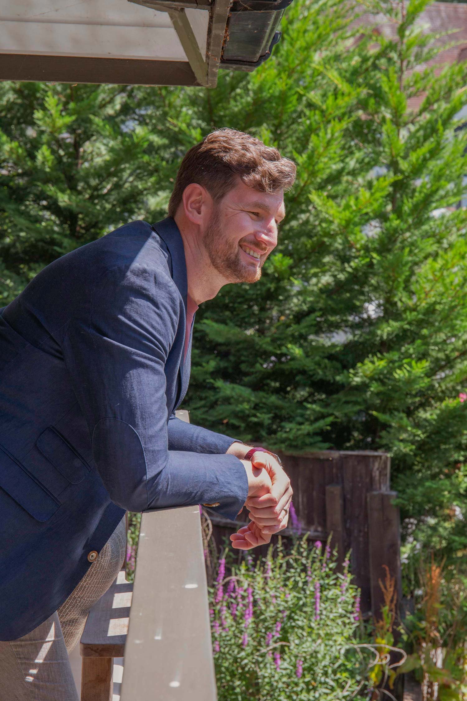 A man with short brown hair and a beard, dressed in a navy blazer and gray pants, smiling and shaking hands outdoors. Behind him are trees and a garden with purple flowers.