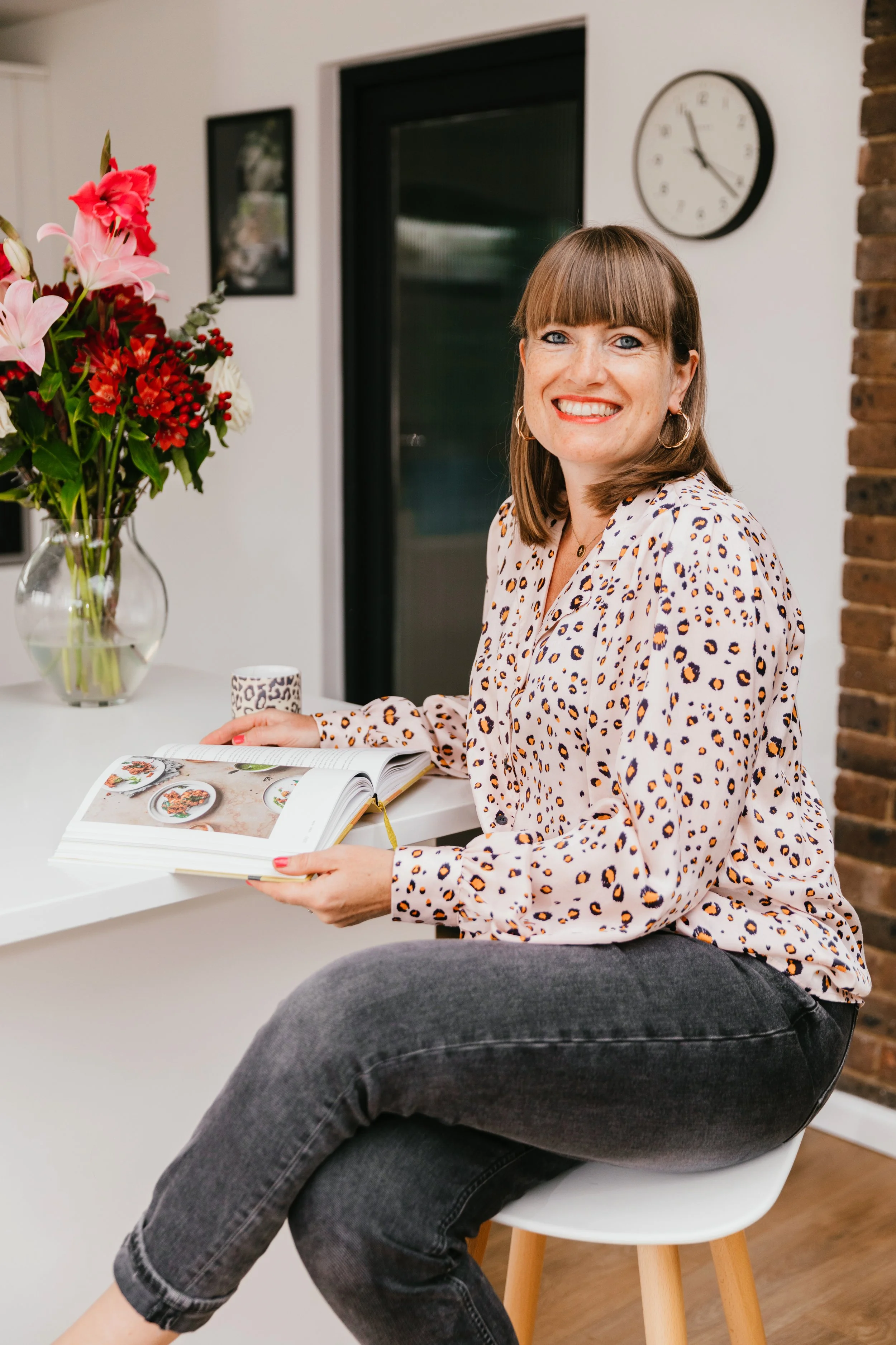 A smiling woman with brown hair and blue eyes sitting at a table with a magazine, in a room with a vase of pink and red flowers, a wall clock, and framed pictures.