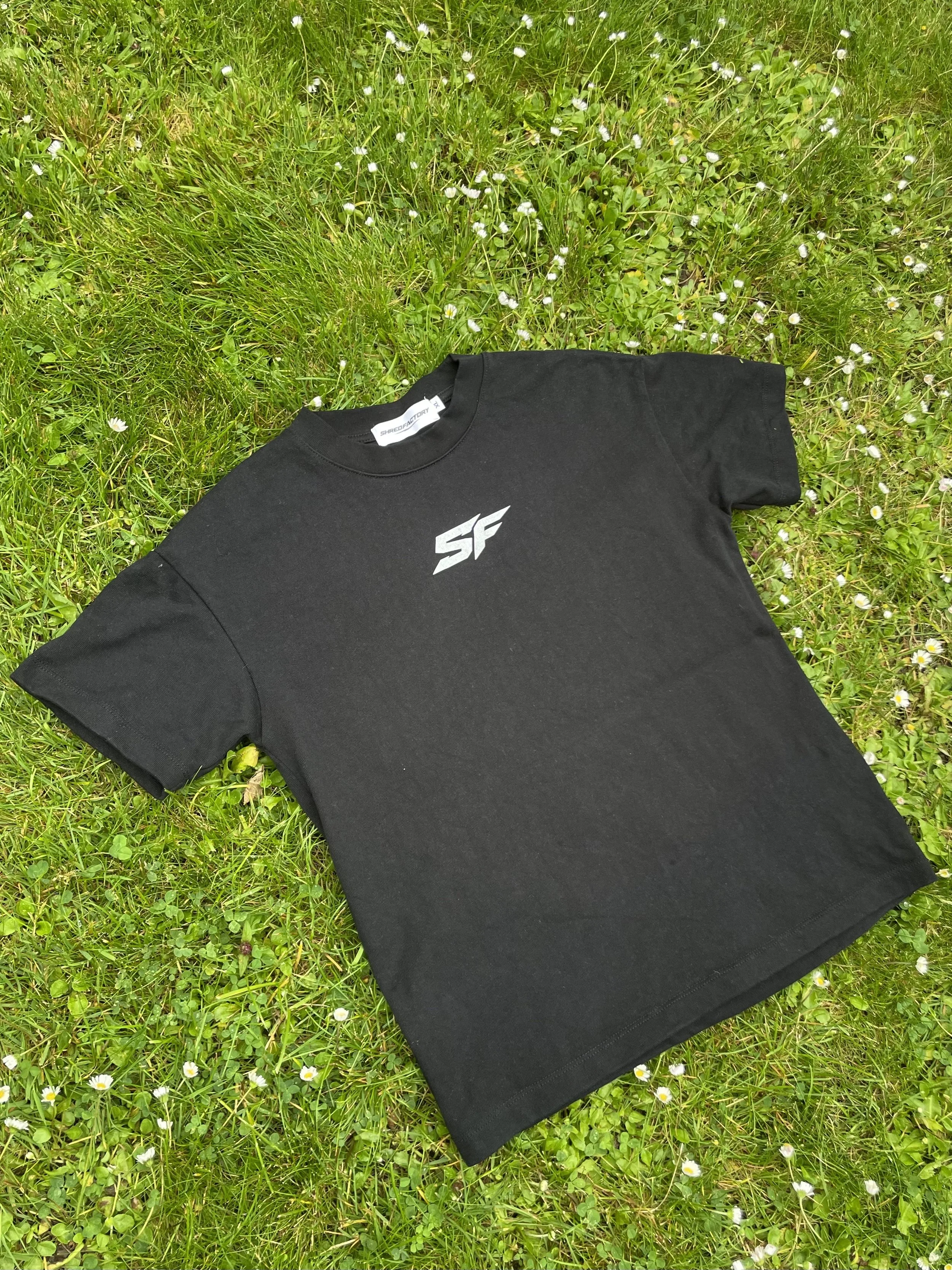 A black T-shirt with a ShredFactory logo lying on green grass with small white flowers.