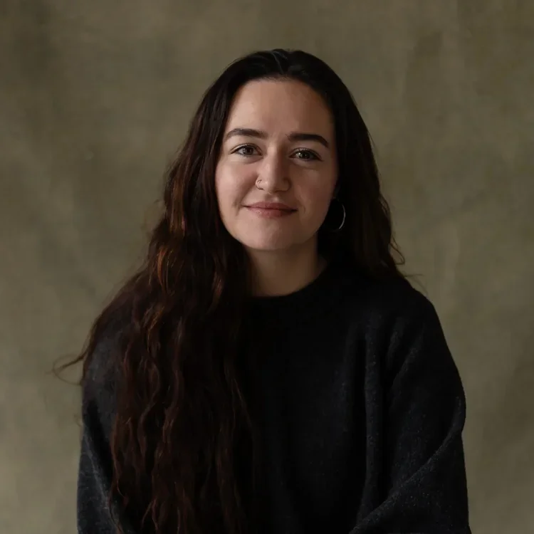 A young woman with long, wavy dark hair, wearing a black sweater, smiling softly against a neutral background.