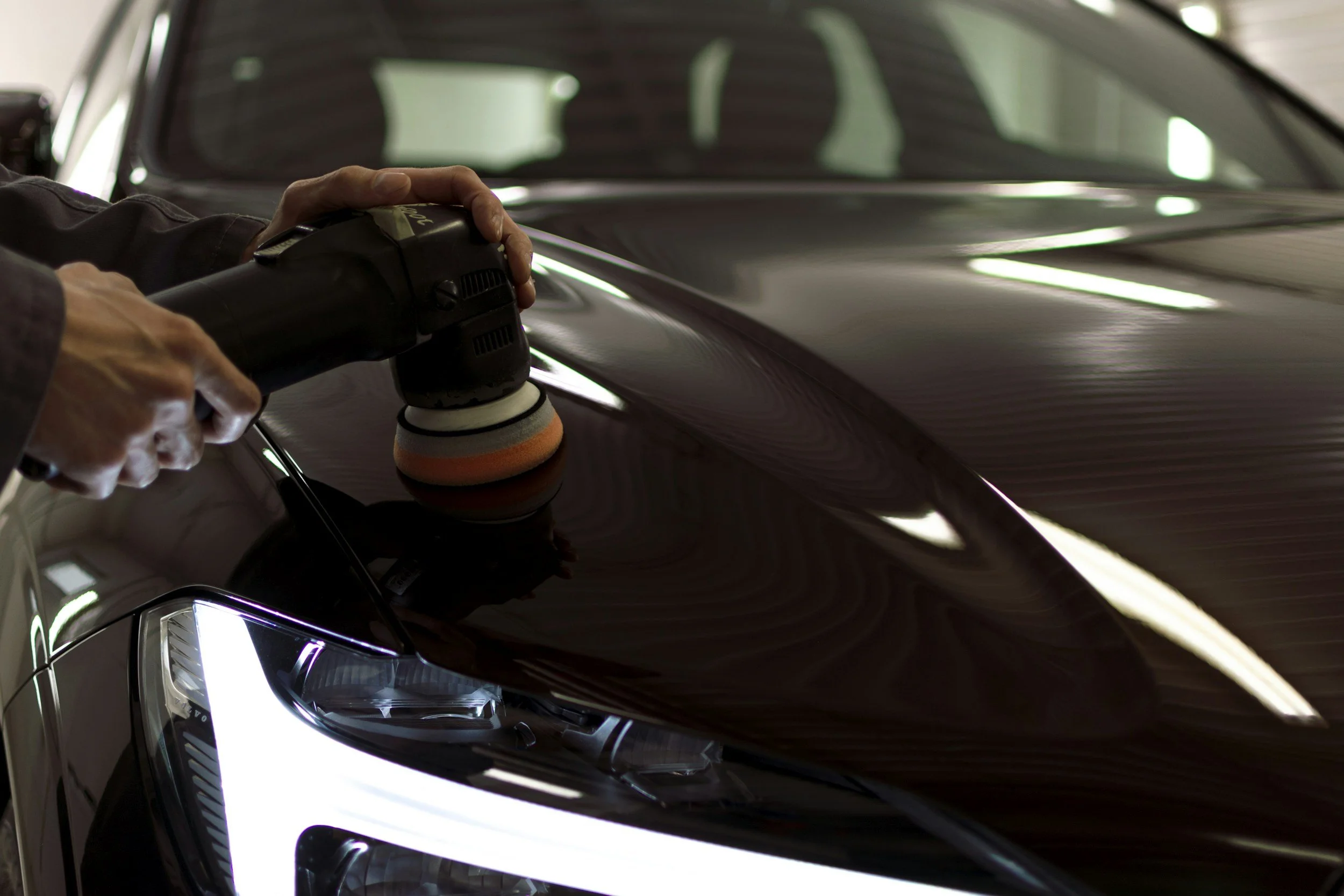 A person polishing a black car with a handheld buffer. The car's front is visible, including the headlight.