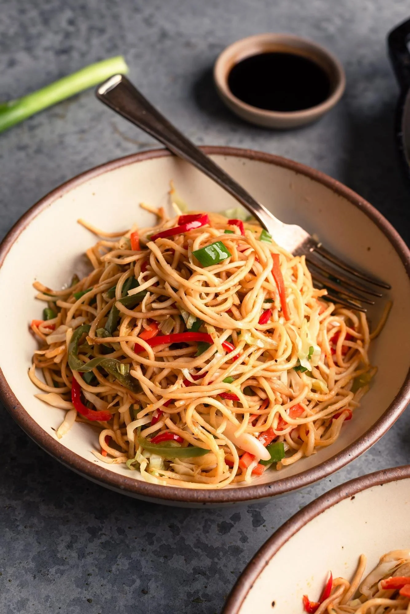 A bowl of Asian-style stir-fried noodles with vegetables, including red bell peppers, green onions, and bean sprouts, on a dark gray surface with a small bowl of soy sauce and chopsticks.