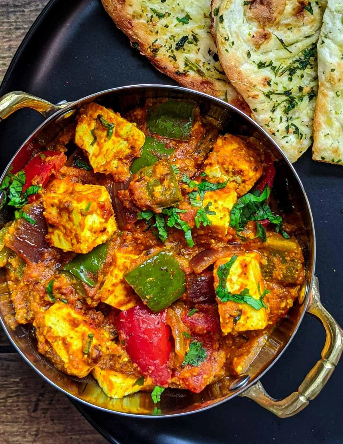 A bowl of Indian-style paneer curry with green peppers, tomato, and spices, garnished with cilantro, placed on a black tray alongside garlic naan bread.