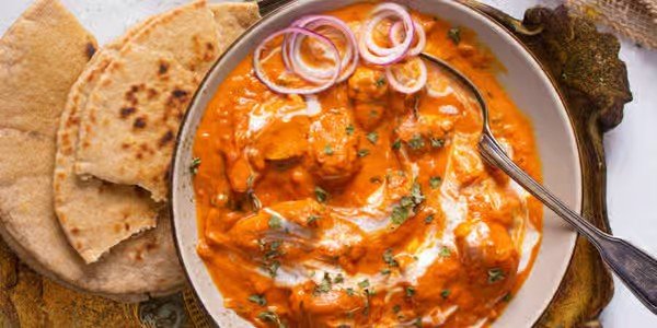 A bowl of Indian butter chicken with green herbs, sliced onions, and naan bread on a plate.