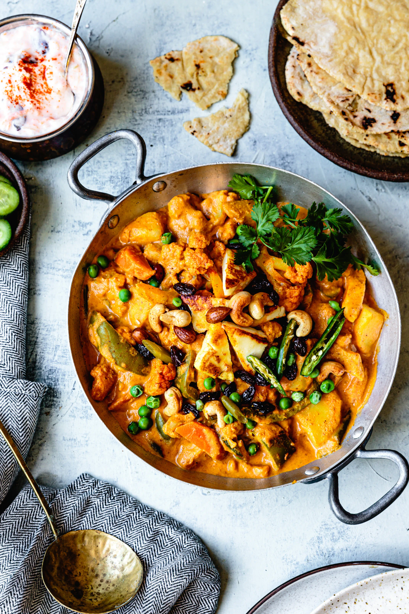A skillet of Indian curry garnished with cilantro, served with naan bread, cucumber slices, and a bowl of yogurt with spices on the side.