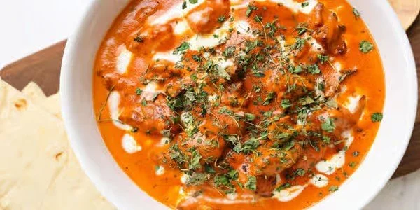 Bowl of Indian butter chicken curry topped with cilantro and cream, served with naan bread on the side.