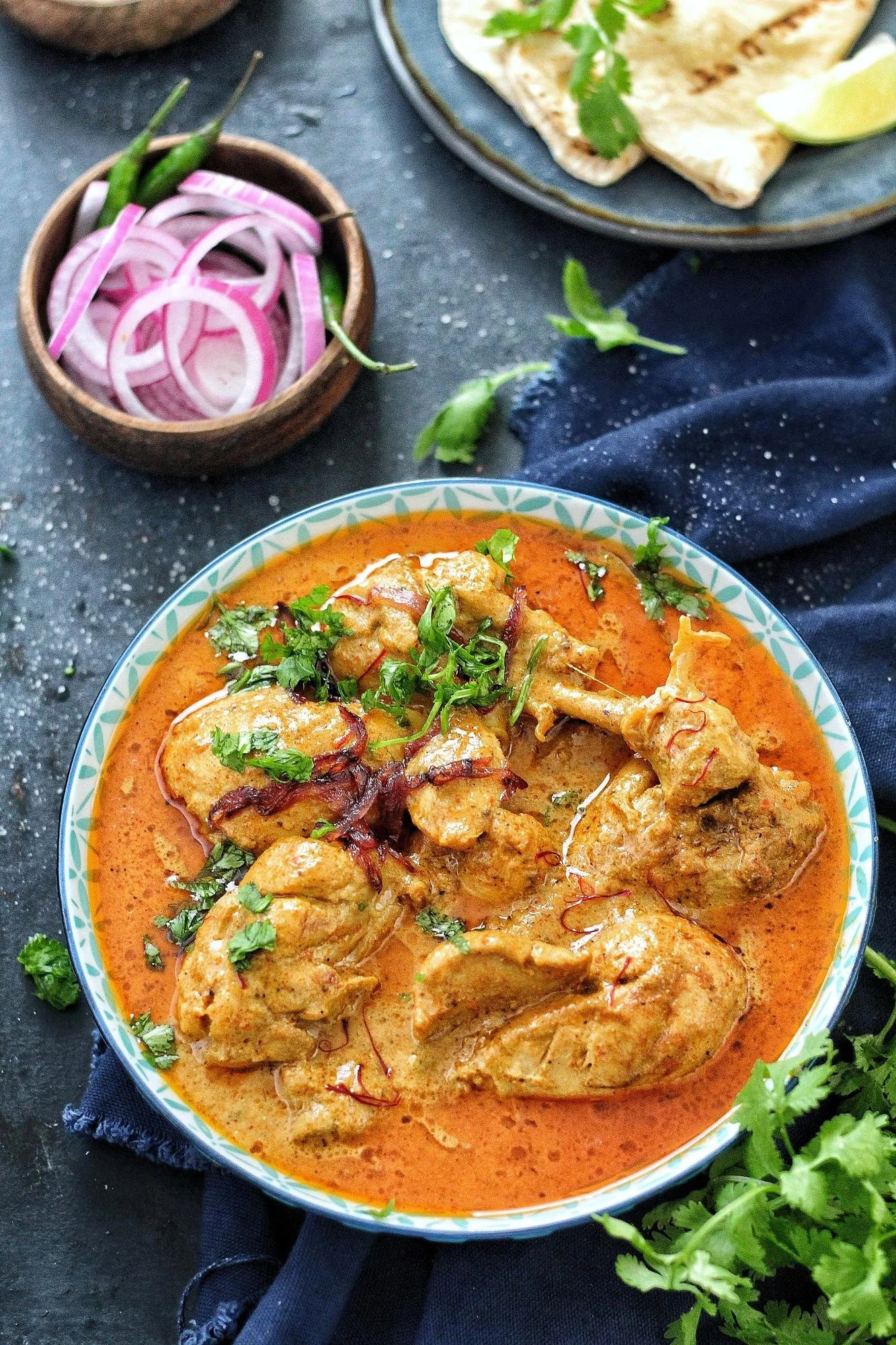 A bowl of chicken curry in orange sauce garnished with cilantro, with a side of sliced onions and green chili peppers in a small brown bowl, and a plate of flatbread with lime wedges in the background.