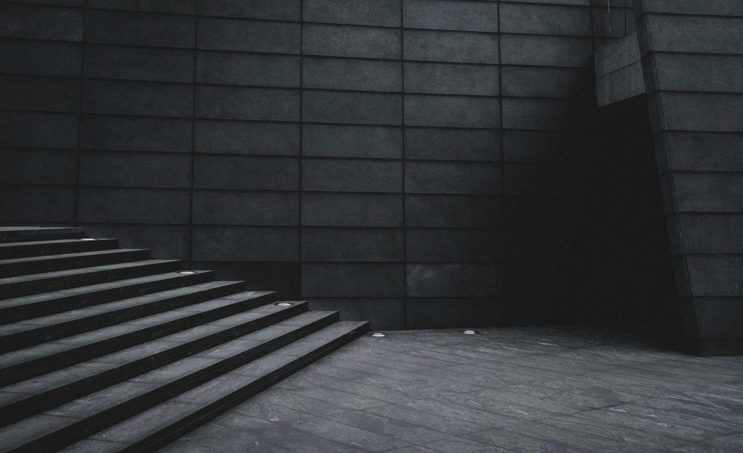 Dark stone staircase with built-in lights at the base of the steps, leading up to a dark stone wall in an industrial or modern building interior.