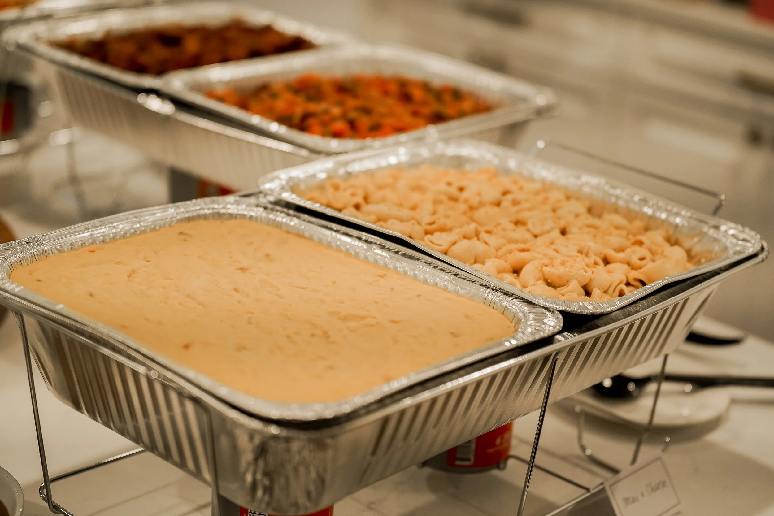 Multiple aluminum trays of food on a buffet table, including a creamy dish, pasta, and a vegetable casserole.