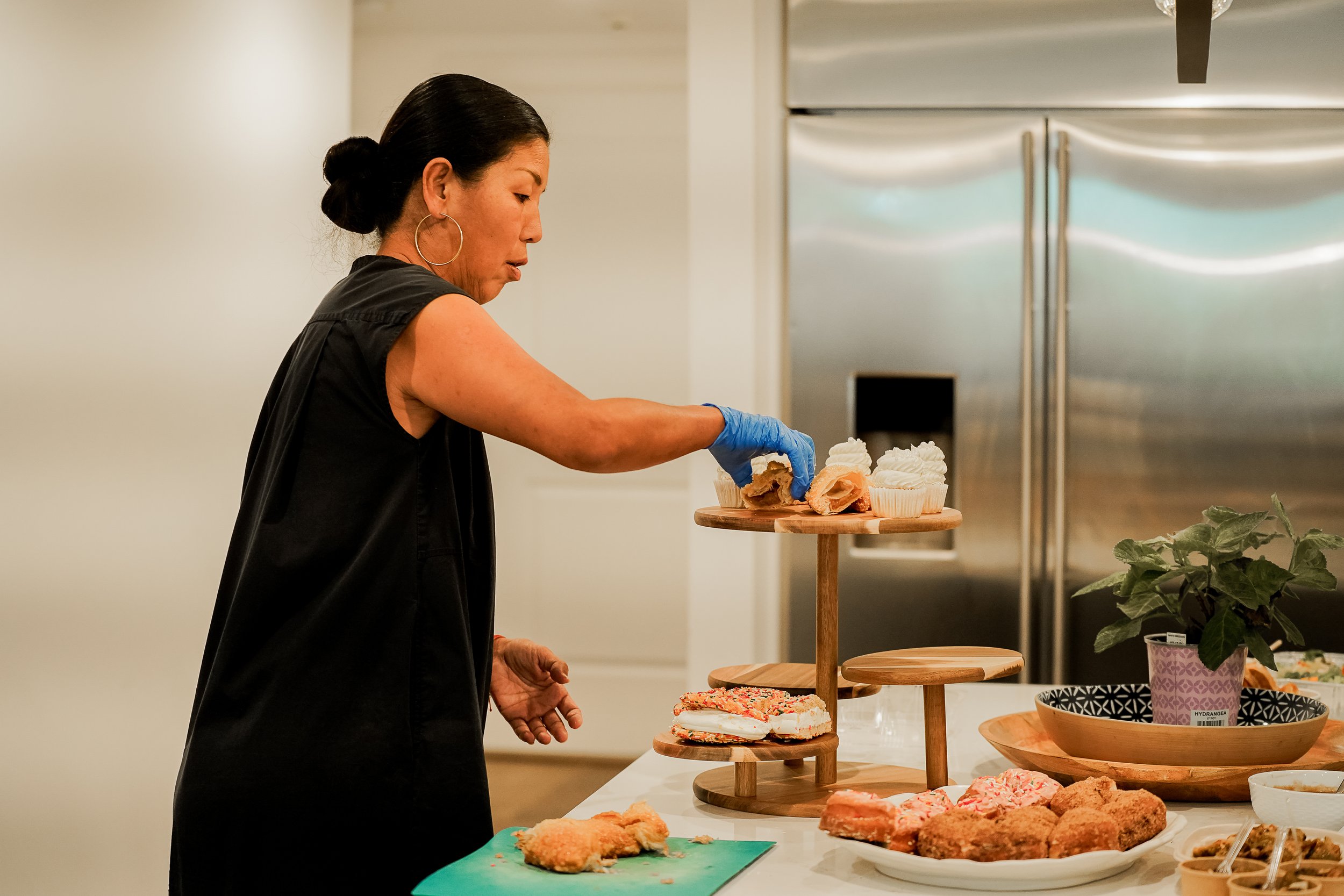 A woman with black hair tied in a bun, wearing large hoop earrings and black clothing, is serving herself food from a buffet table. The table has various dishes including sandwiches, fried foods, cupcakes, and other snacks, with a tiered wooden serving stand.