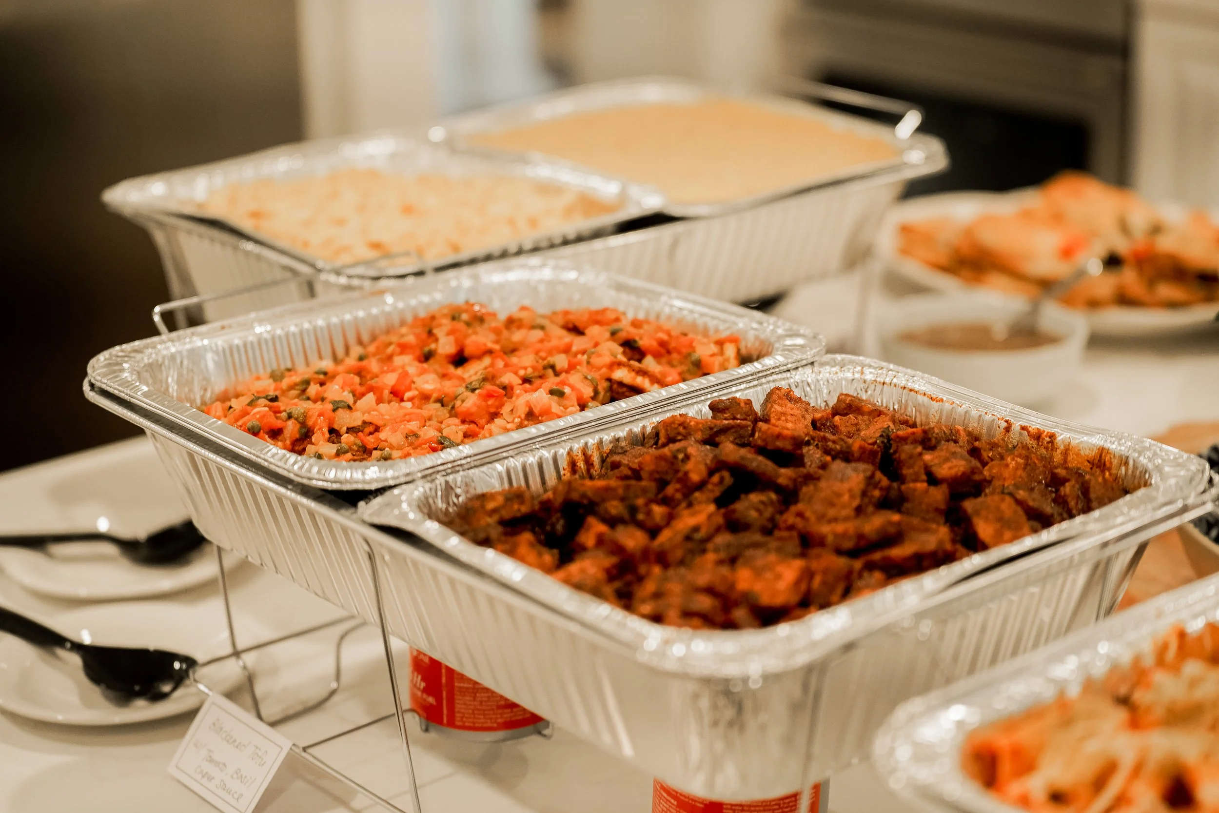 Various trays of food, including potato salad, chicken, and other dishes, on a table for a buffet or gathering.
