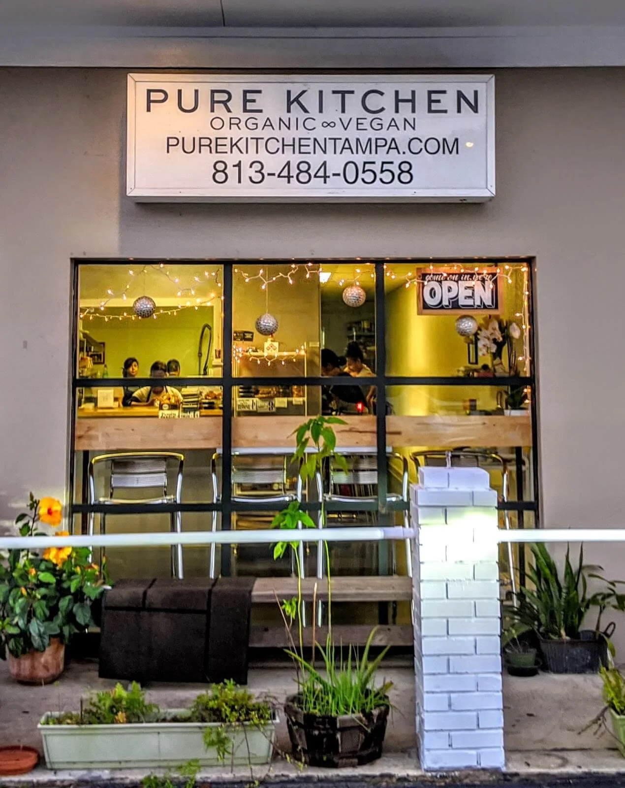 The exterior of a small restaurant named Pure Kitchen, decorated with string lights and ornaments, with a visible sign that reads 'Open.' Through the window, customers can be seen inside, and outside there are potted plants and a small garden area.