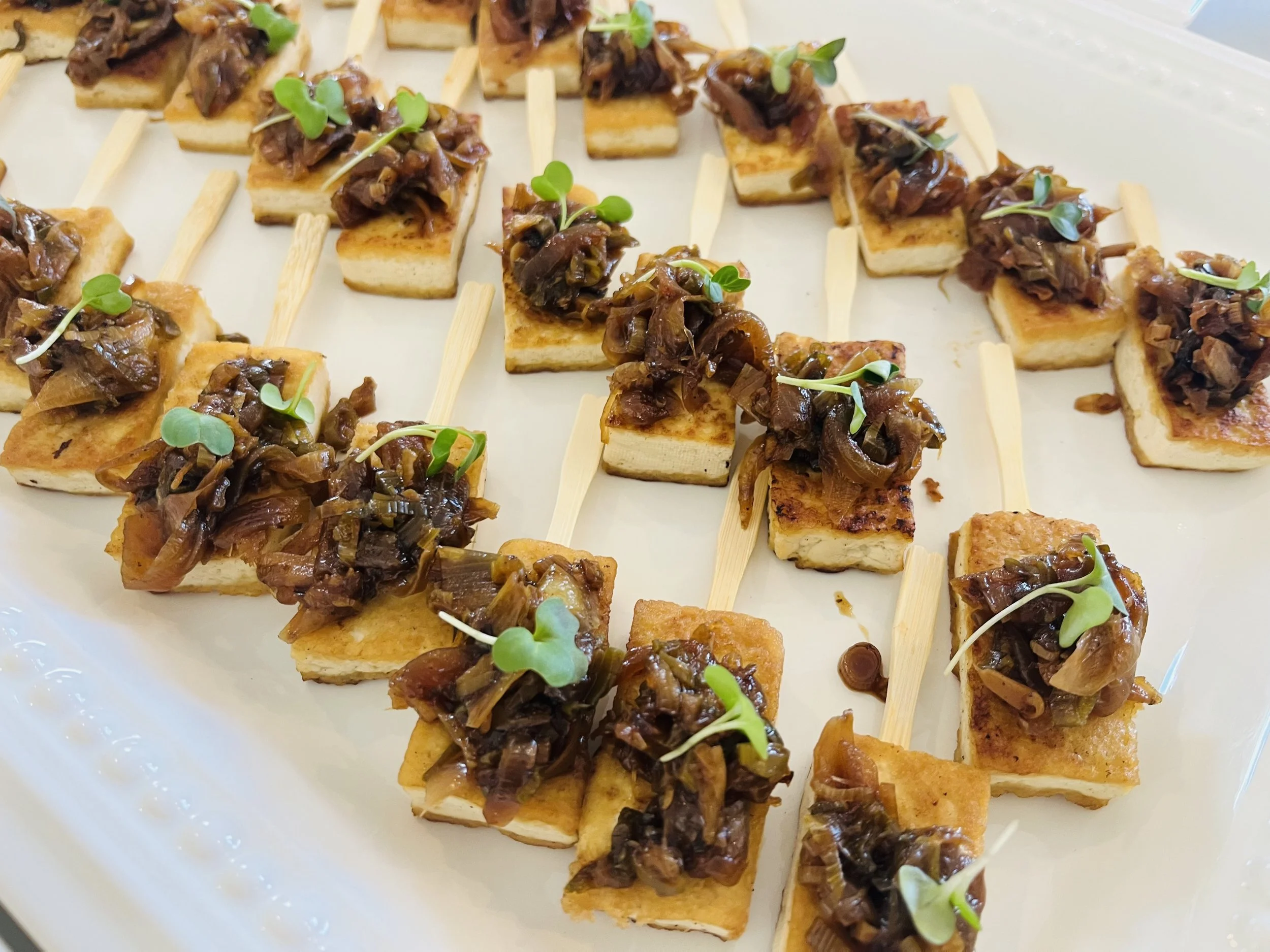 Several small rectangular pieces of toasted bread topped with caramelized onions and microgreens on a white platter.
