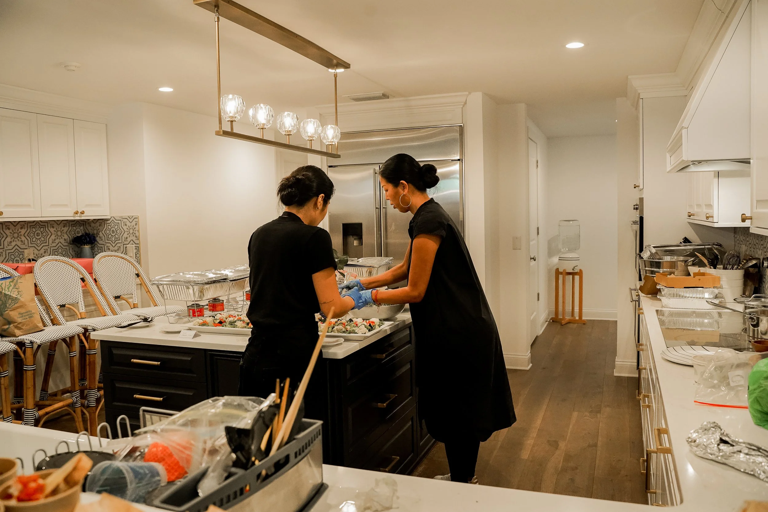 Two women preparing food in a kitchen with white cabinets, a stainless steel refrigerator, and pendant lighting, surrounded by various kitchen supplies and dishes.
