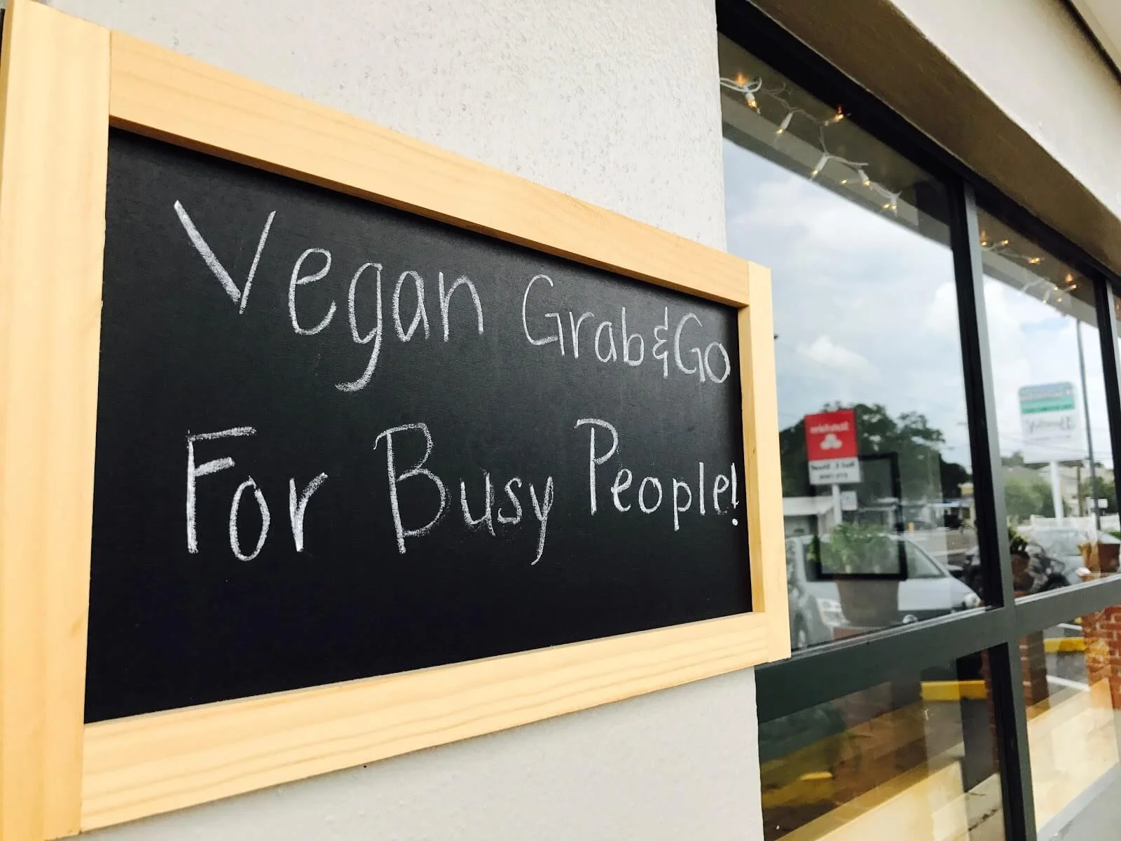 A black chalkboard sign with a wooden frame that reads 'Vegan Grab & Go For Busy People!' in white chalk, outside a store with large glass windows reflecting the parking lot and cloudy sky.