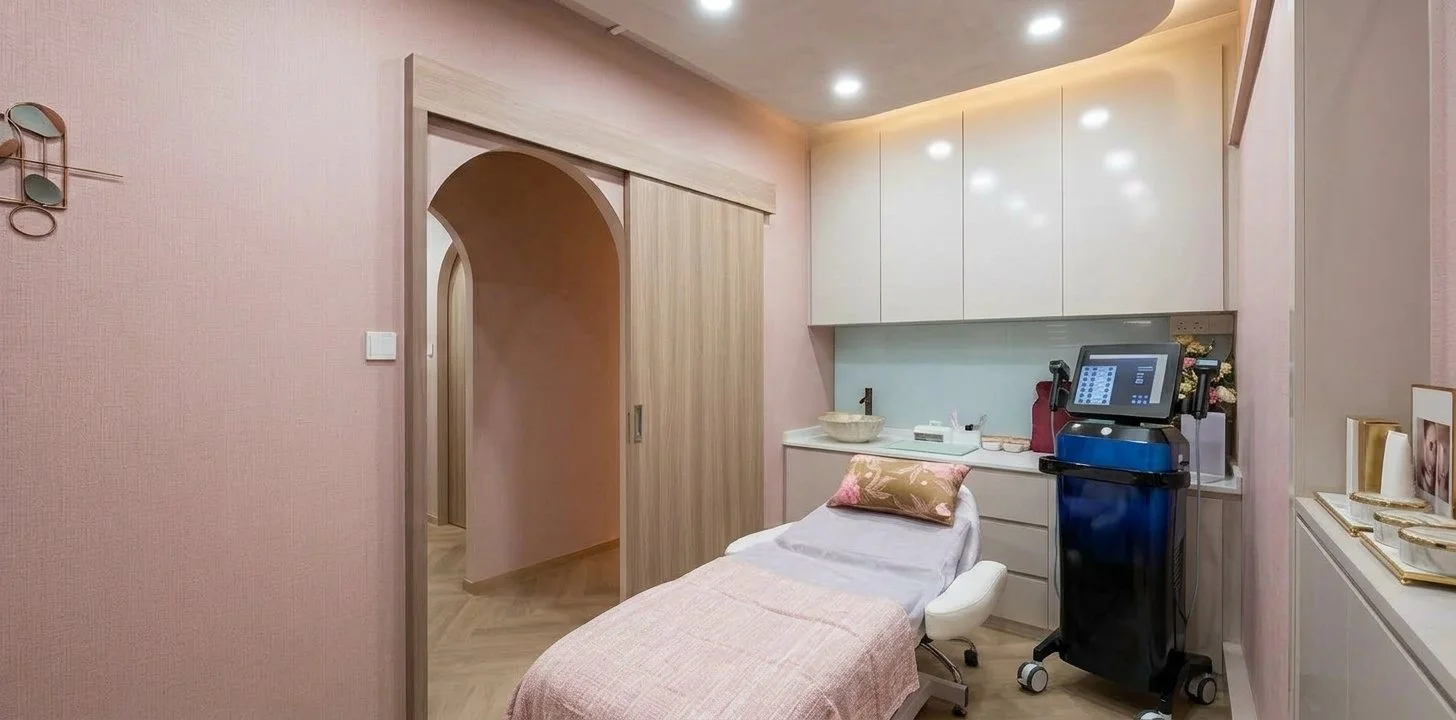 A cozy room with pink textured walls, a treatment or examination bed covered with a pink blanket, and a decorative pillow. There are white cabinets above and below a small countertop with medical or skincare equipment. A computer or touchscreen display is mounted on a medical device on a cart. The room has warm lighting and a hallway with an arched doorway leading to another area.