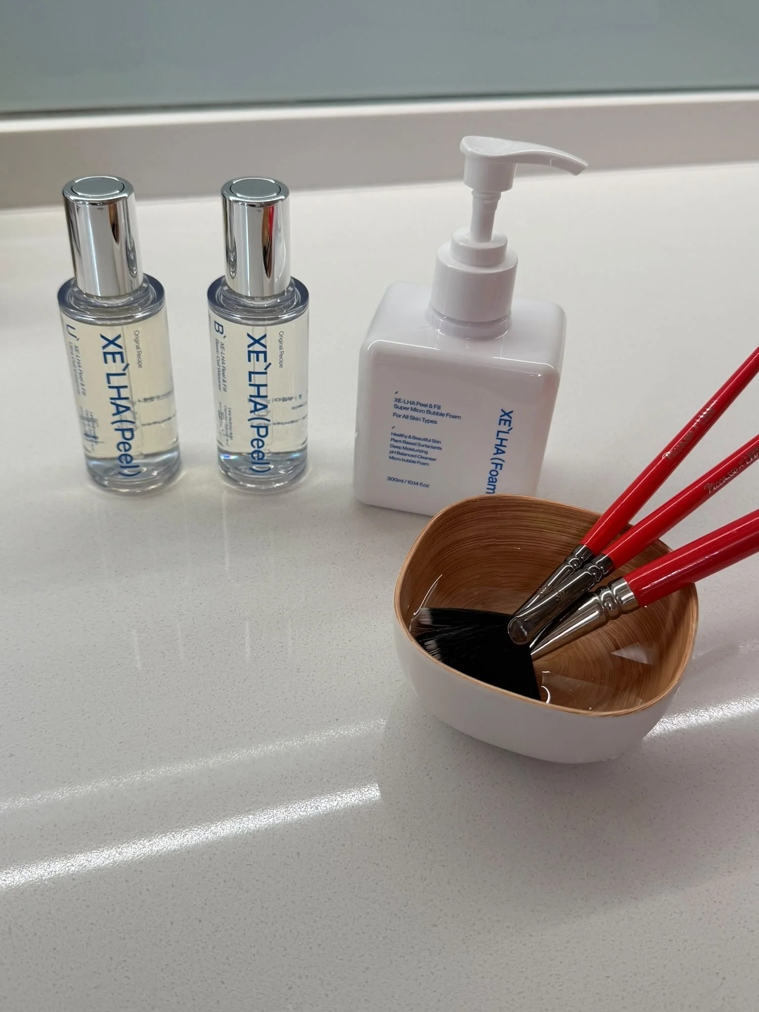 Skincare products including a pump bottle of foam cleanser, two small bottles of skincare serum, and a small bowl with makeup brushes on a white surface.