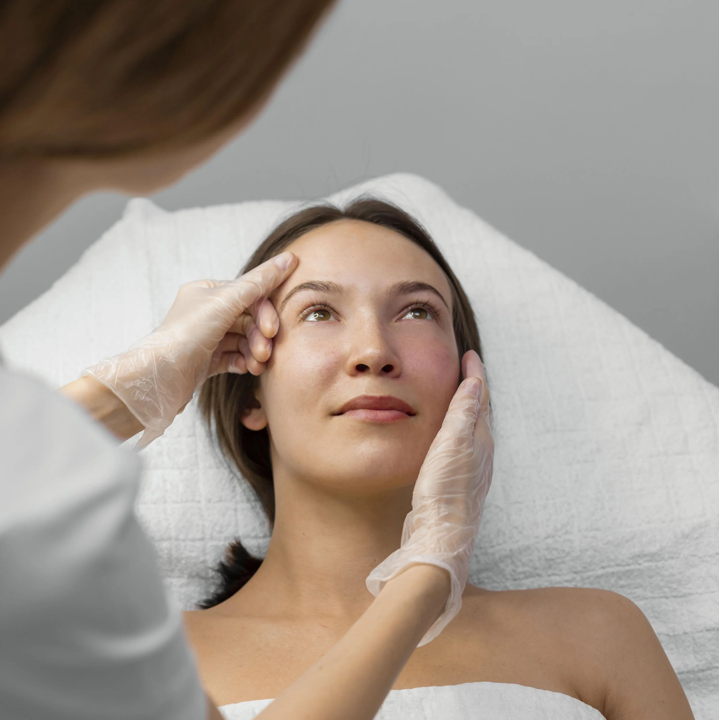 A woman lying on a medical examination bed, getting her eyebrows examined or treated by a healthcare professional wearing gloves.