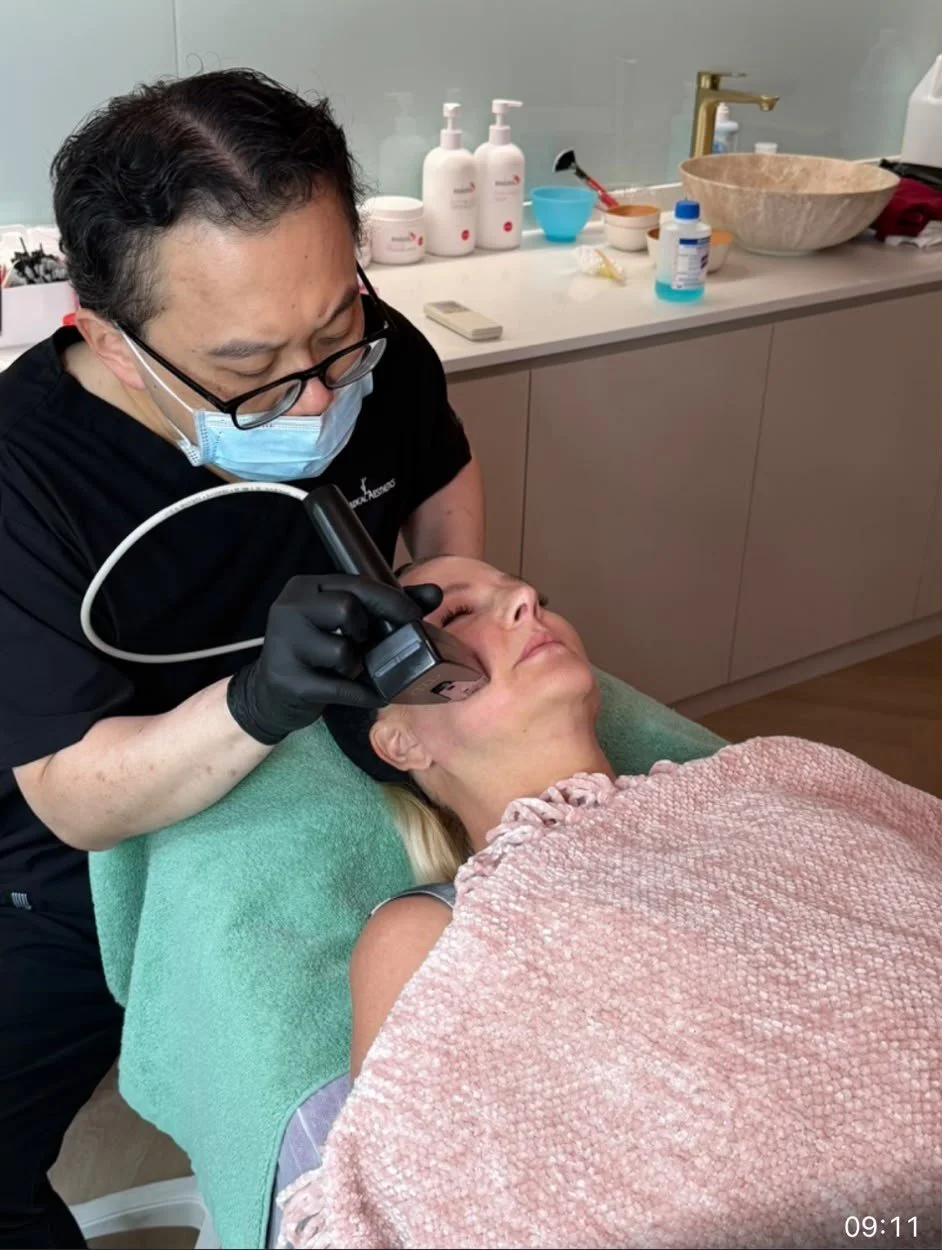 A woman receiving a cosmetic treatment on her face from a professional in a clinic or spa setting. The practitioner is using a handheld device on the woman's face, and both are wearing masks.