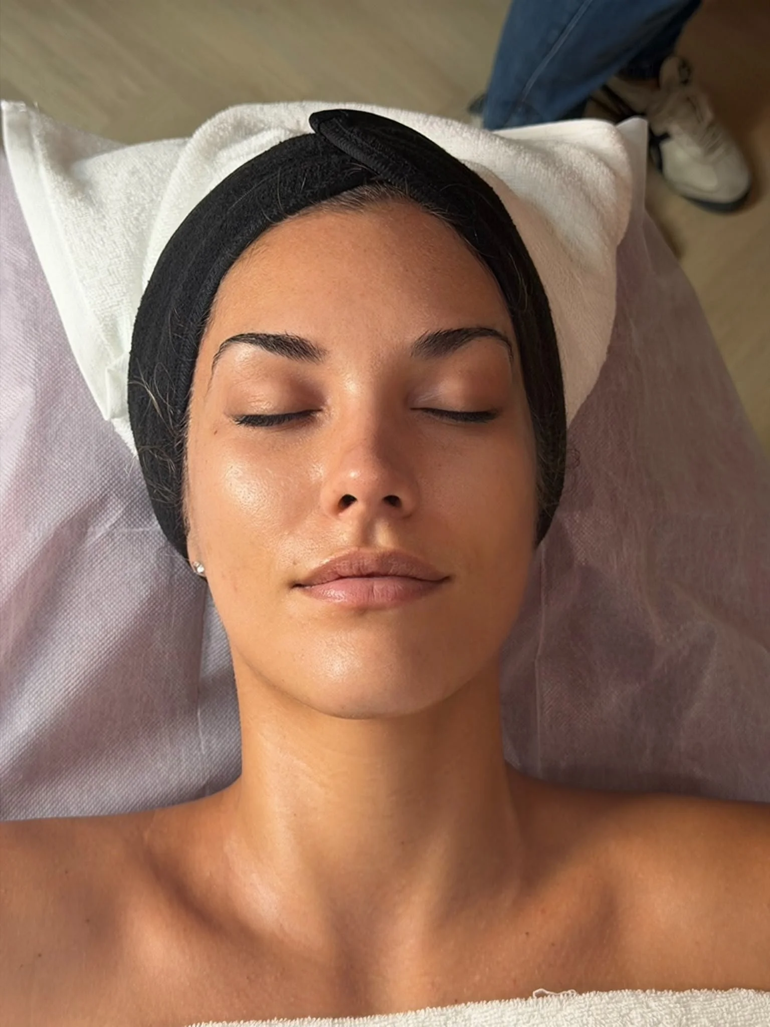Close-up of a woman with closed eyes and a relaxed expression, lying on a treatment bed with her head resting on a white towel, wearing a black headband, in a spa or beauty clinic setting.