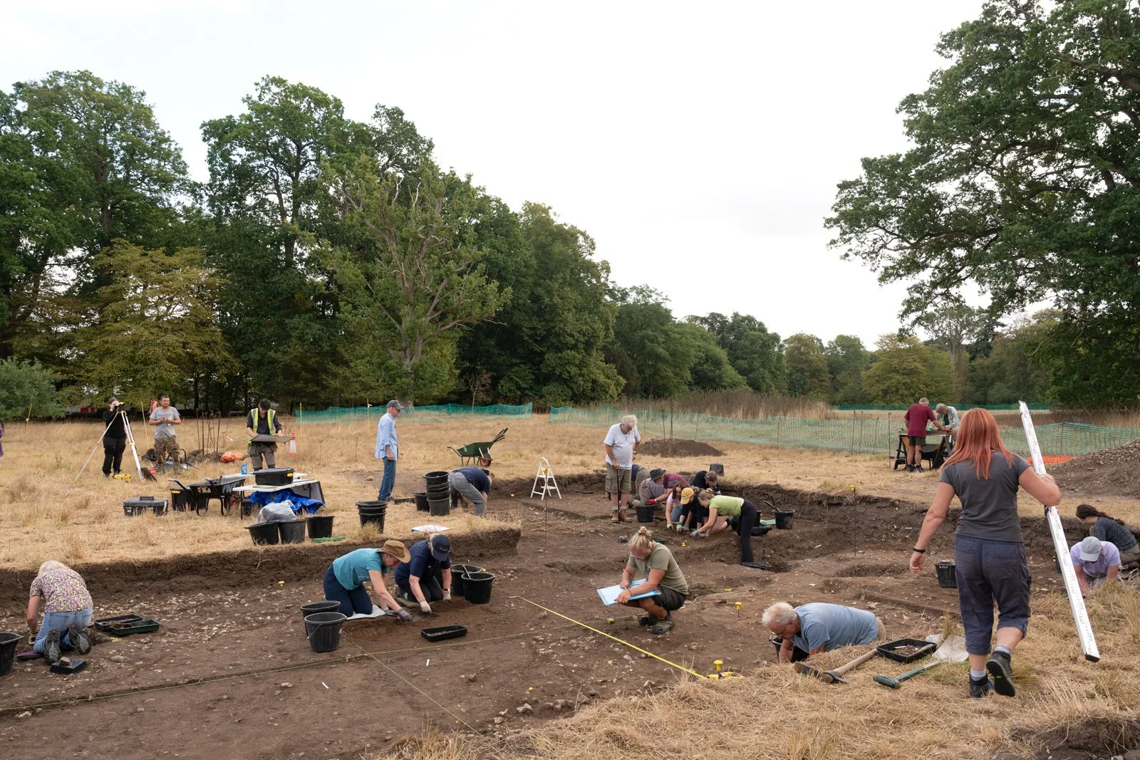 Archaeologists at an excavation site in a grassy field surrounded by large trees, working together to uncover artifacts.