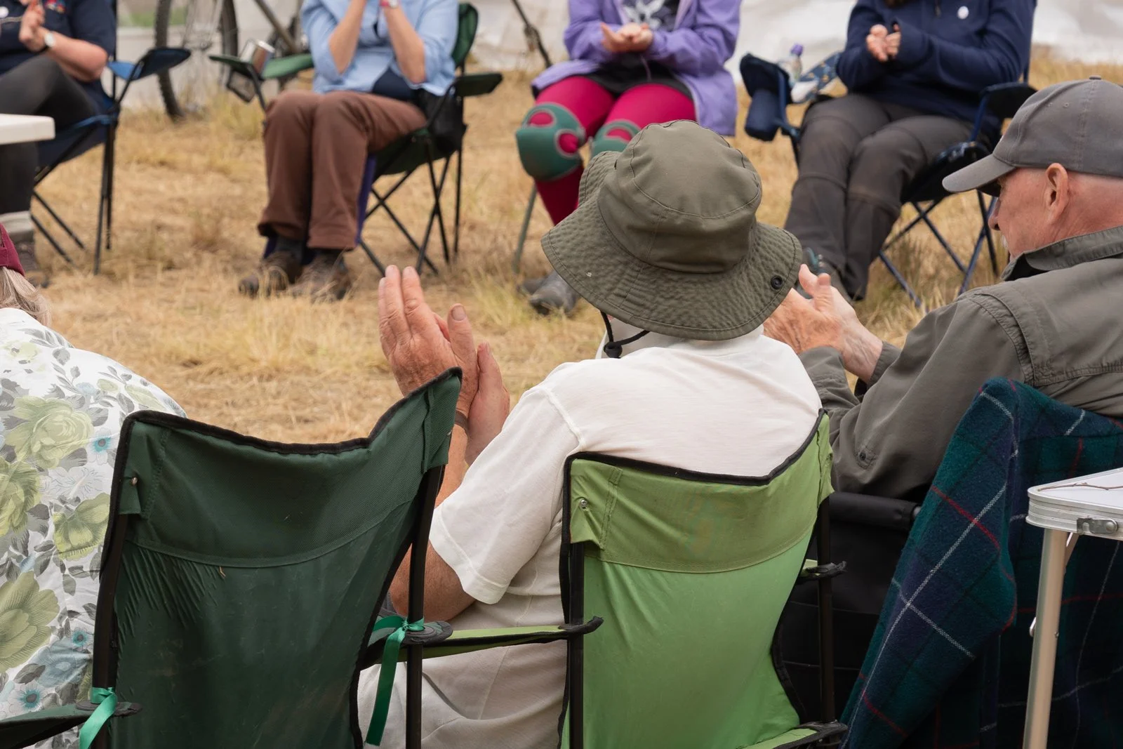 Group of elderly people sitting outdoors in a semi-circle, engaged in a conversation or activity, with grass and trees in the background.