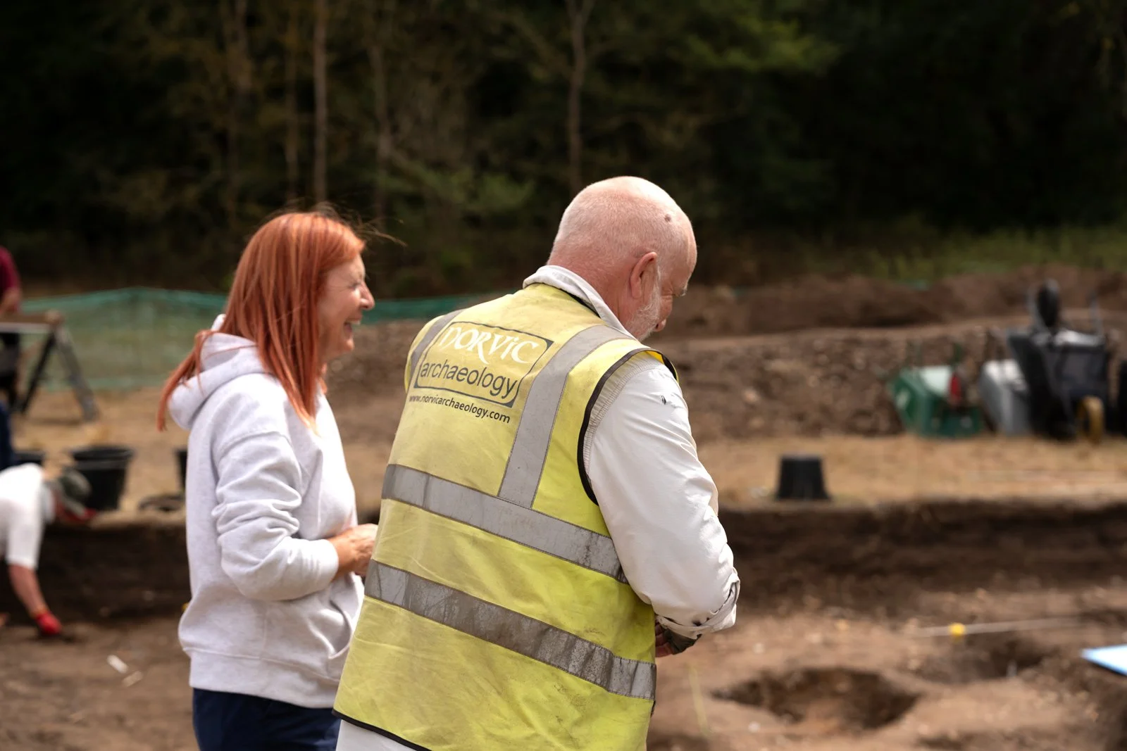 Two people, a woman with red hair and a man with a shaved head wearing a yellow archaeological vest, standing at an outdoor excavation site with trees in the background.