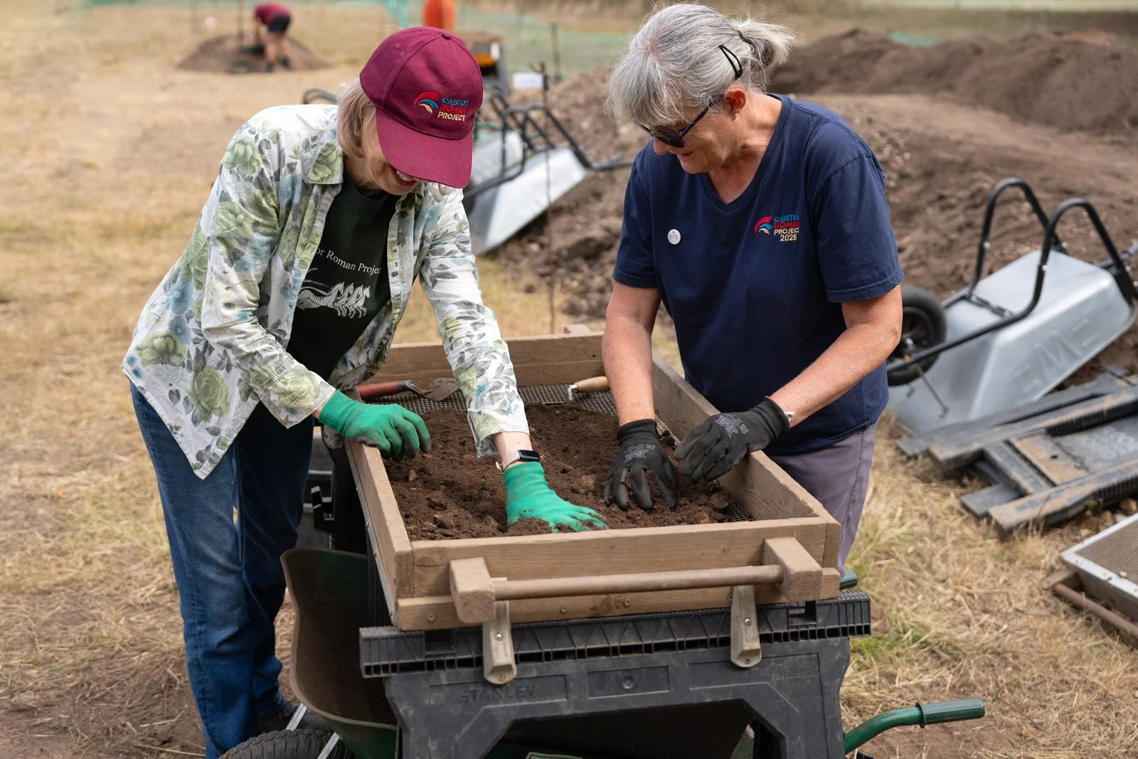 Two women working together planting in a garden bed outdoors, wearing gloves and casual clothing, with gardening tools and supplies nearby.