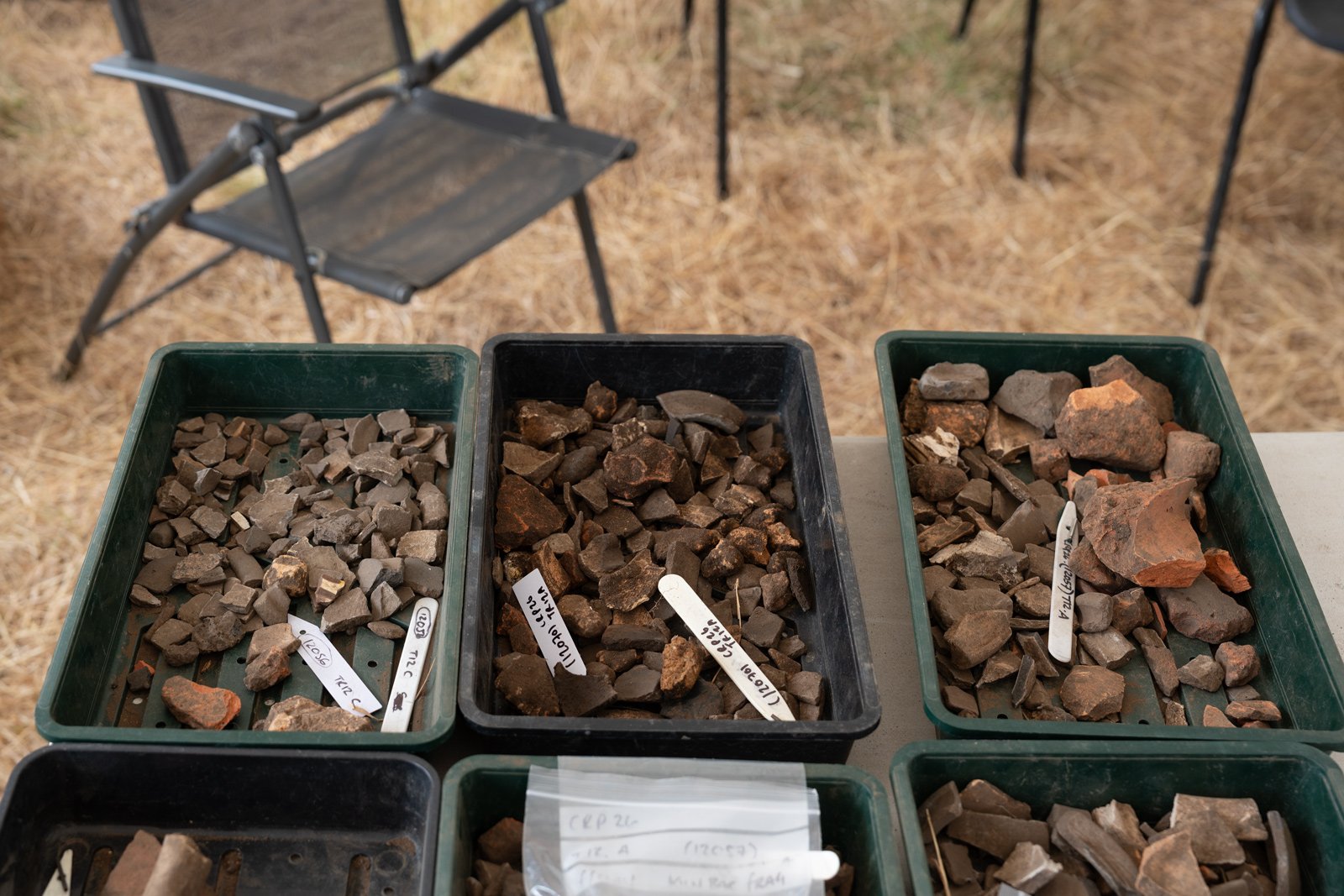 Plastic trays filled with various types of rocks and minerals, labeled with handwritten tags, set on a table outdoors with a camping chair and dry grass in the background.