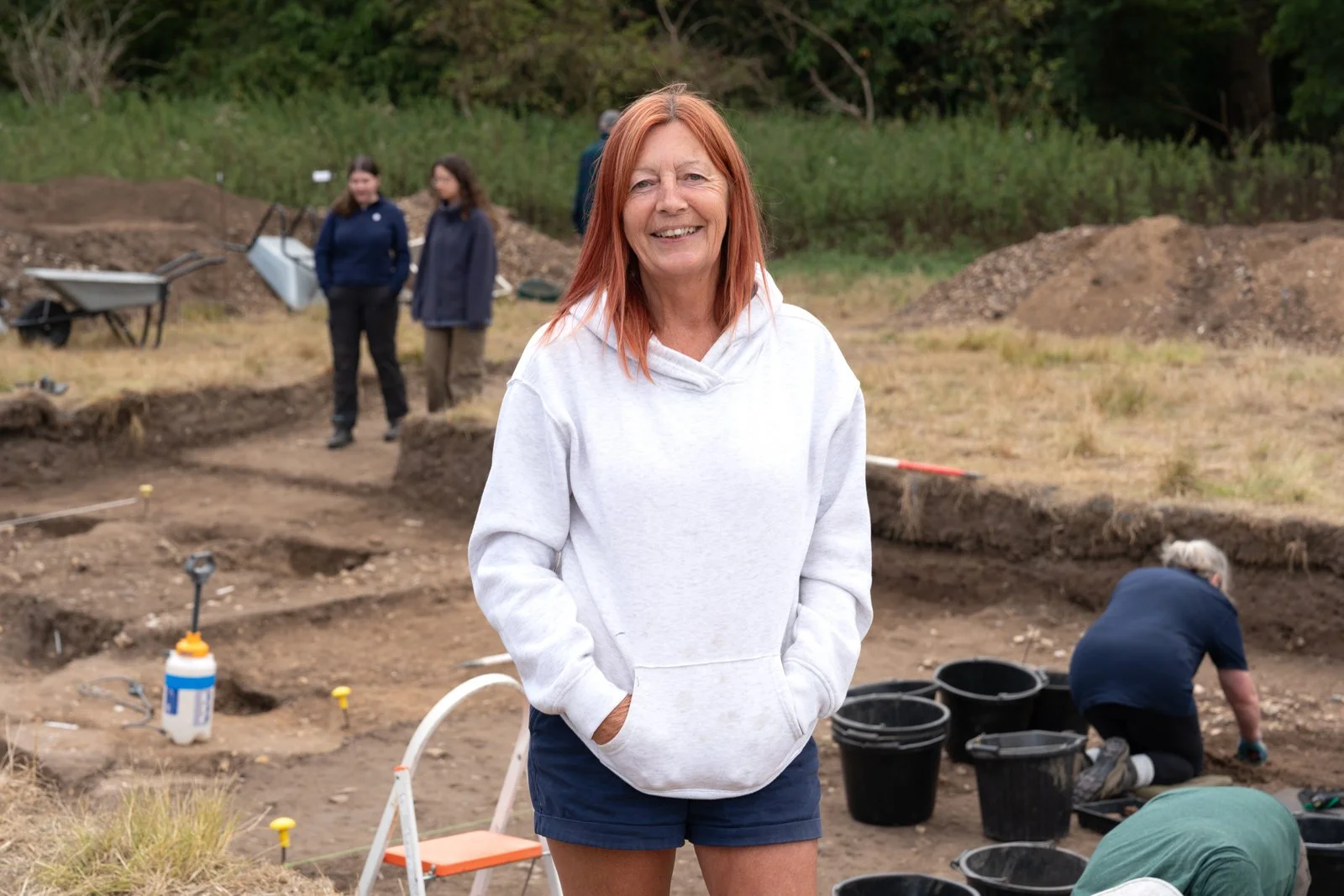 A woman with red hair wearing a light gray hoodie and shorts, smiling at the camera, standing outdoors at an archaeological excavation site with buckets and tools, and people working in the background.