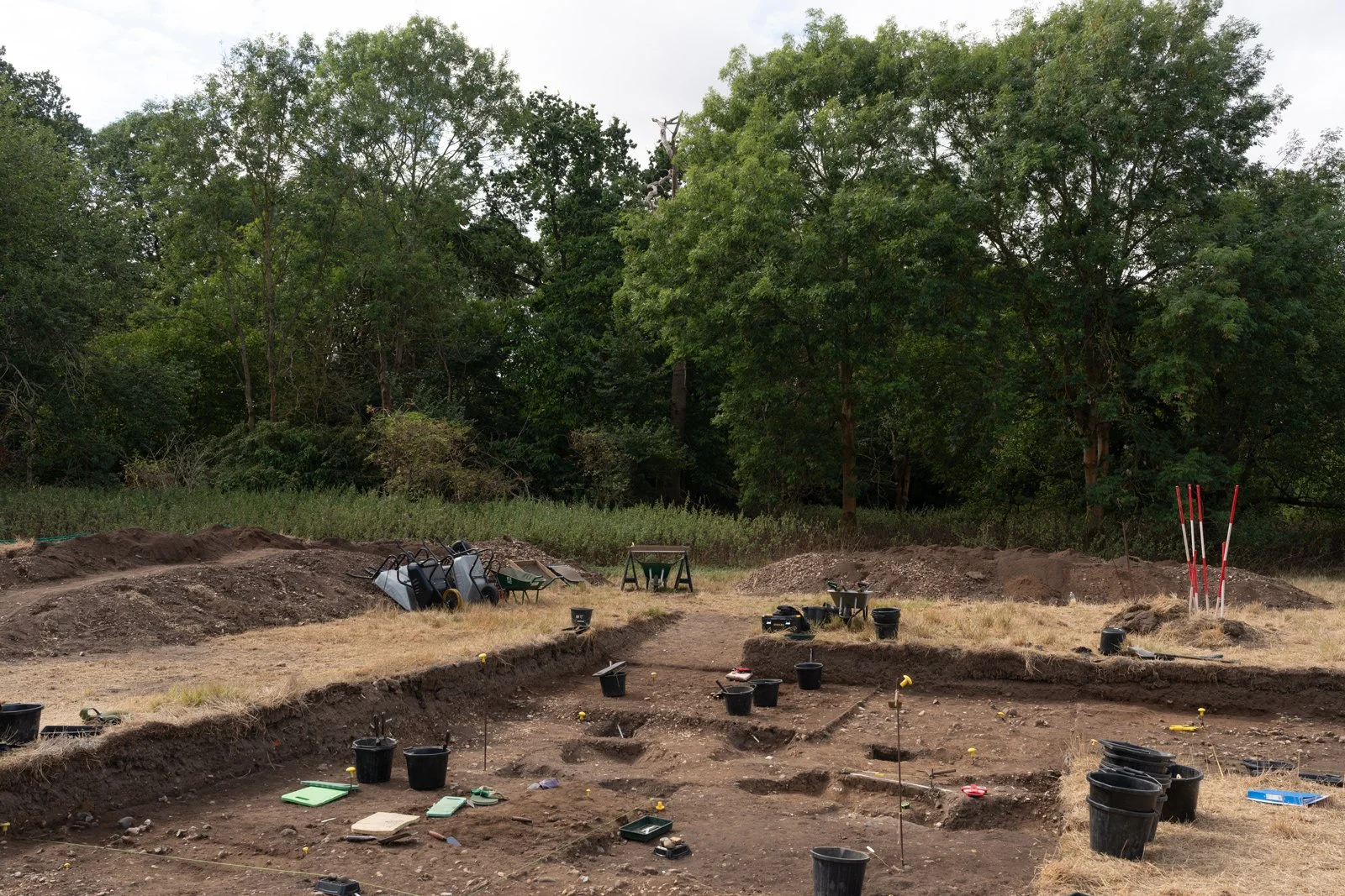 Archaeological excavation site with trenches, tools, and buckets, surrounded by trees.