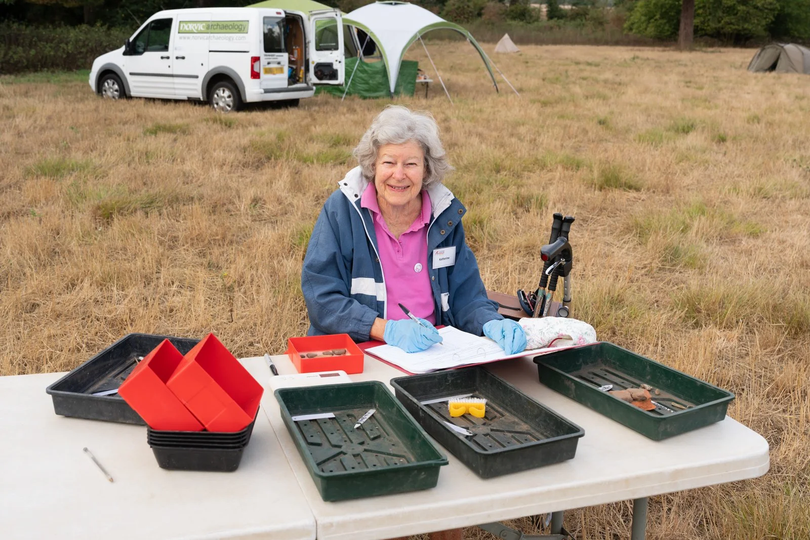 A woman with gray hair, wearing a pink shirt, blue jacket, and blue gloves, sits at a white table outdoors on a grassy field, working with excavation tools. Behind her are tents and a white van with the logo "Nonvavic Archaeology".