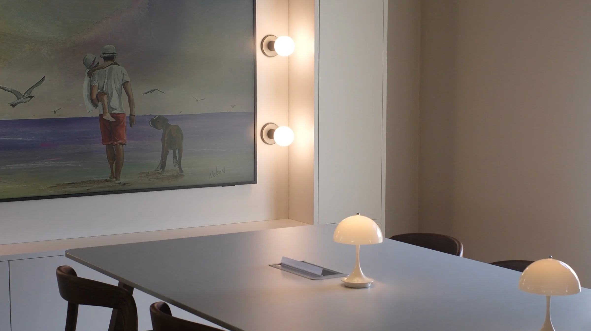 Interior of a room with a modern white wall, decorated with a colorful painting of two children at the beach, and two round wall-mounted lights. There is a conference table with a lamp and chairs around it.
