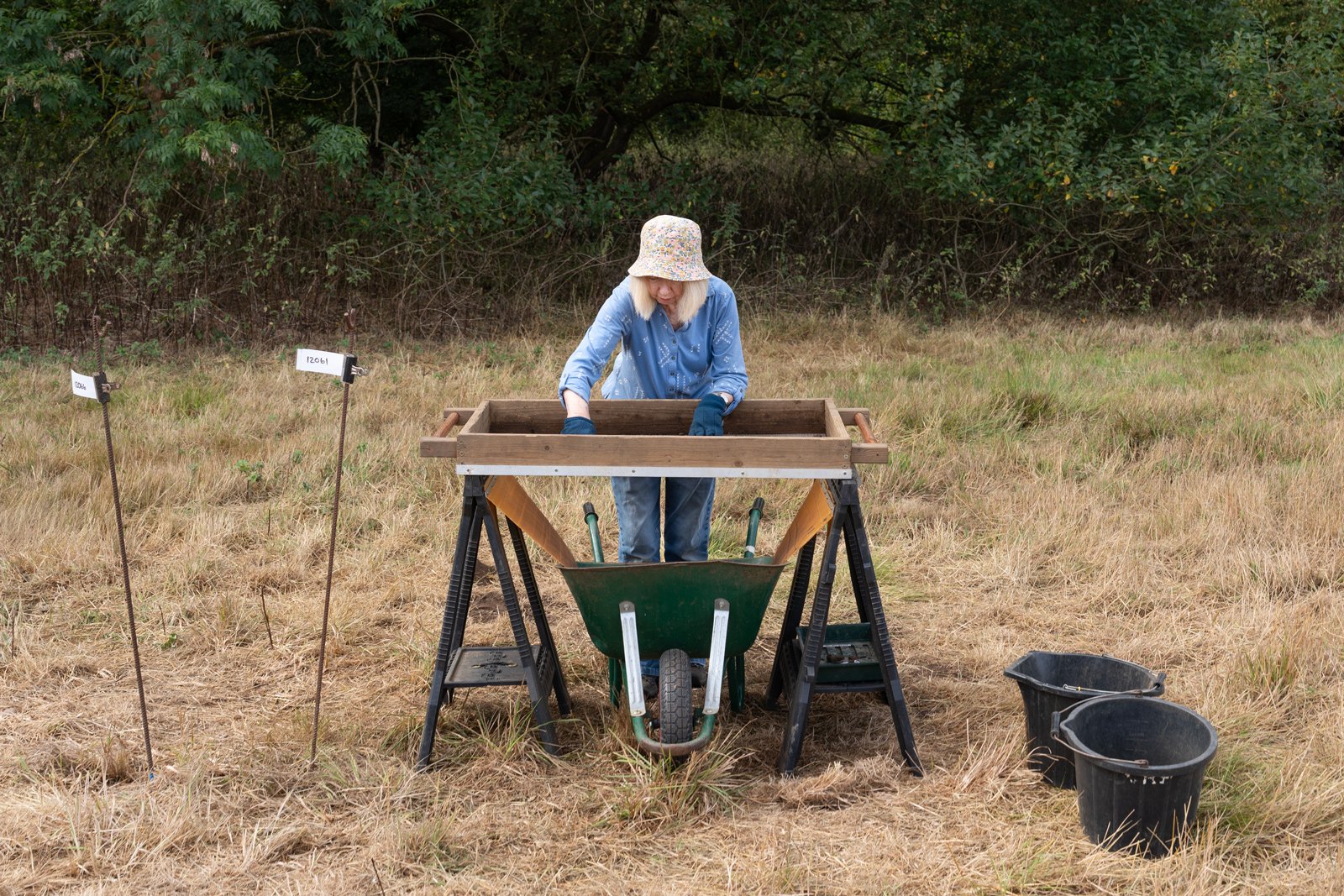 A woman in a blue shirt and floral hat working in a field with a wheelbarrow, black buckets, trees, and grass.