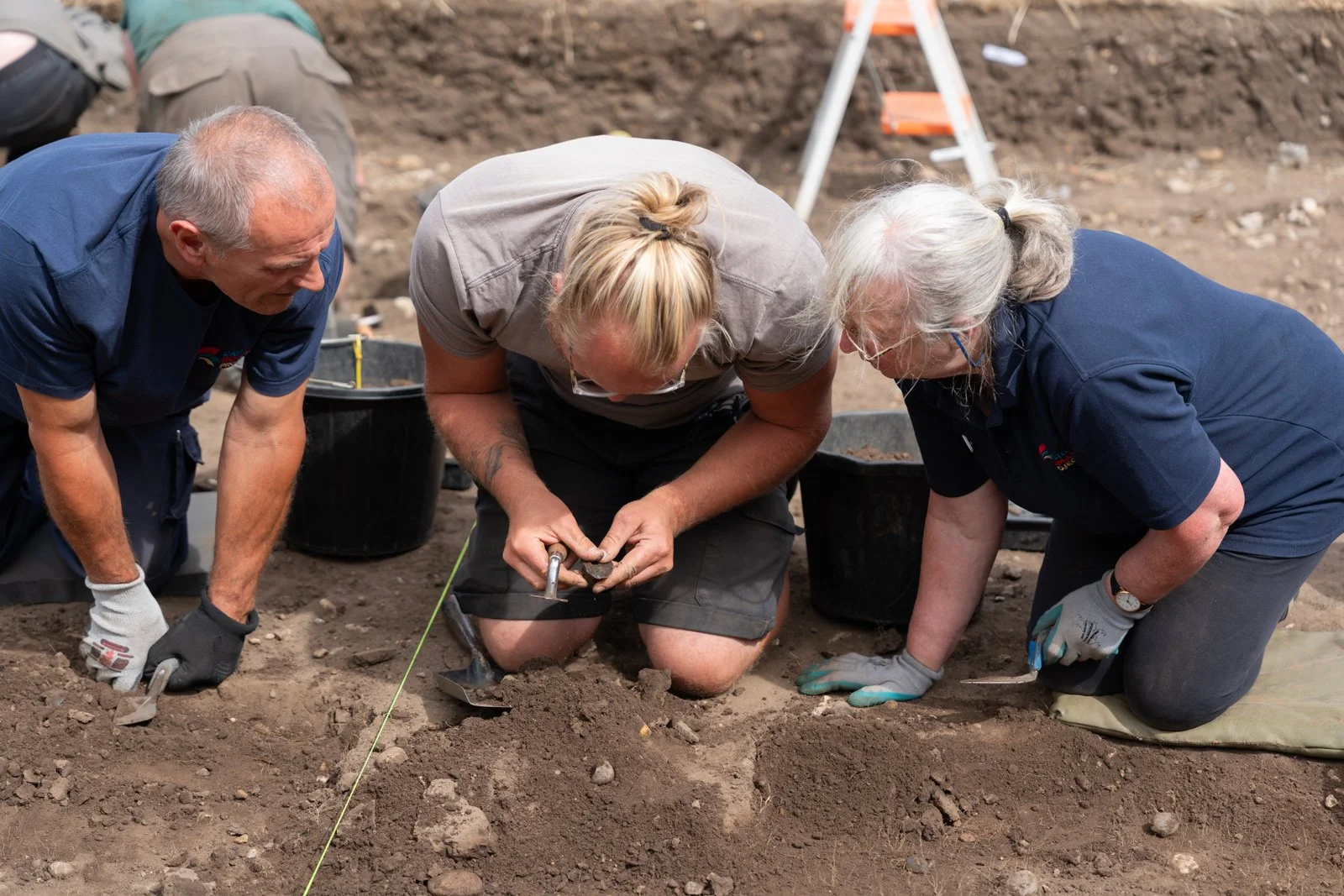Three people working on an archaeological dig, kneeling on the ground and carefully excavating soil with tools.