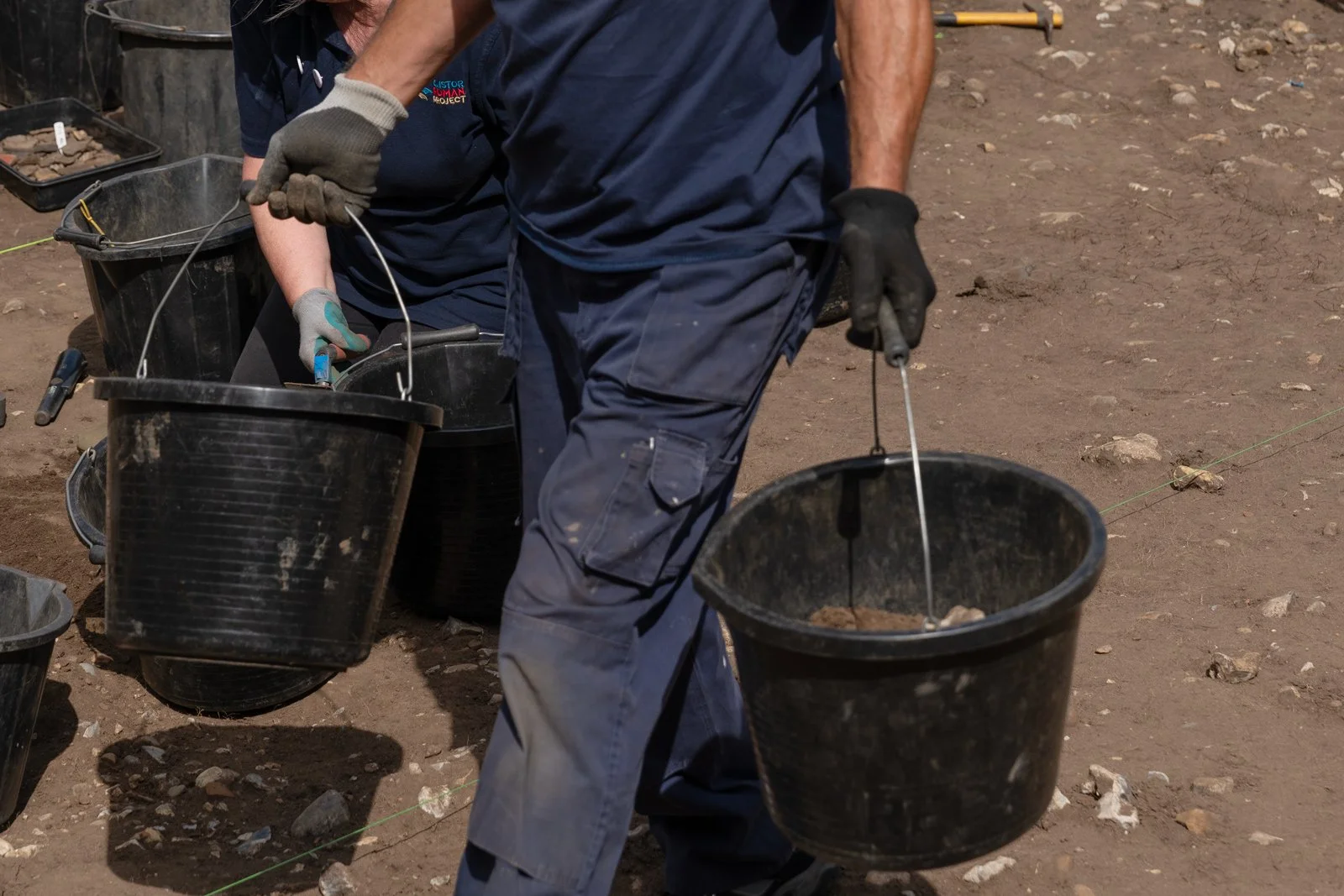People working outdoors with black buckets, tools, and gloves on a dirt surface.