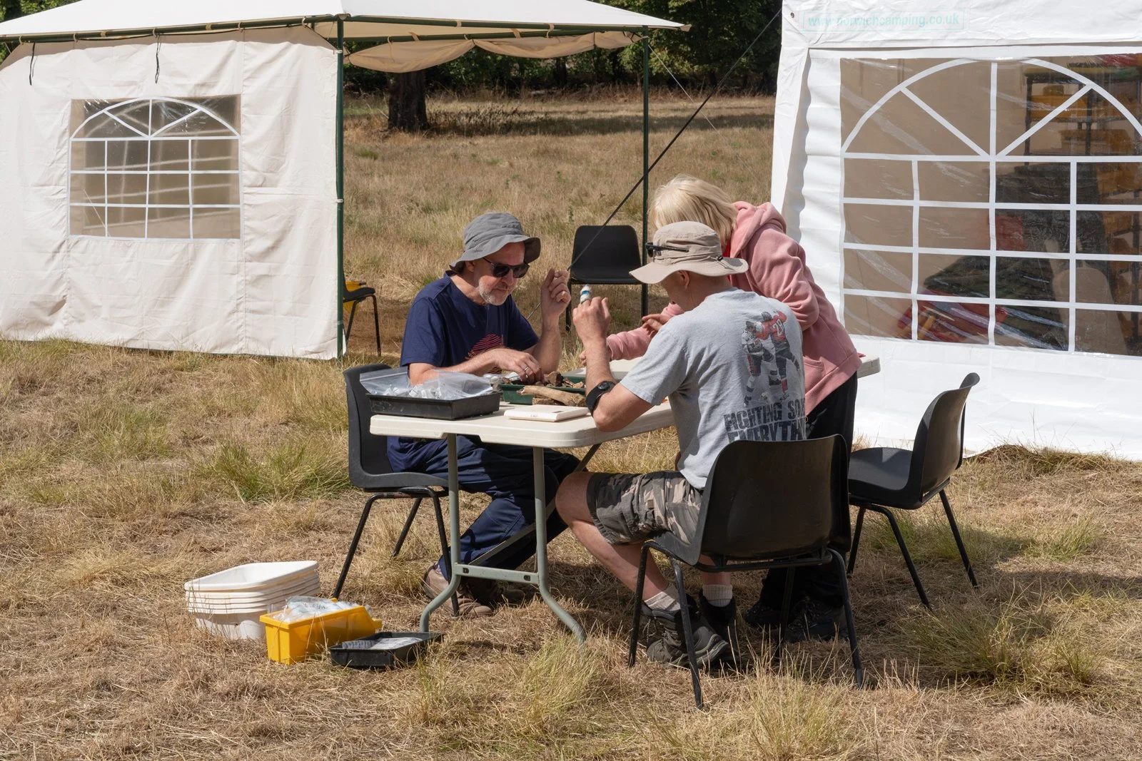 Three people sitting and working at a table outdoors during the daytime. They are under a canopy, with one person wearing a pink hoodie, another with a gray hat and gray t-shirt, and the third with dark sunglasses and a blue t-shirt. The table has va