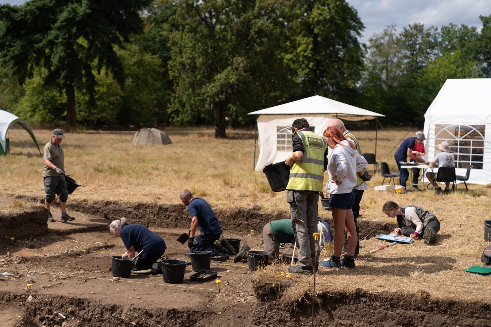 People working and studying outdoors at an archaeological excavation site, with tents and trees in the background.