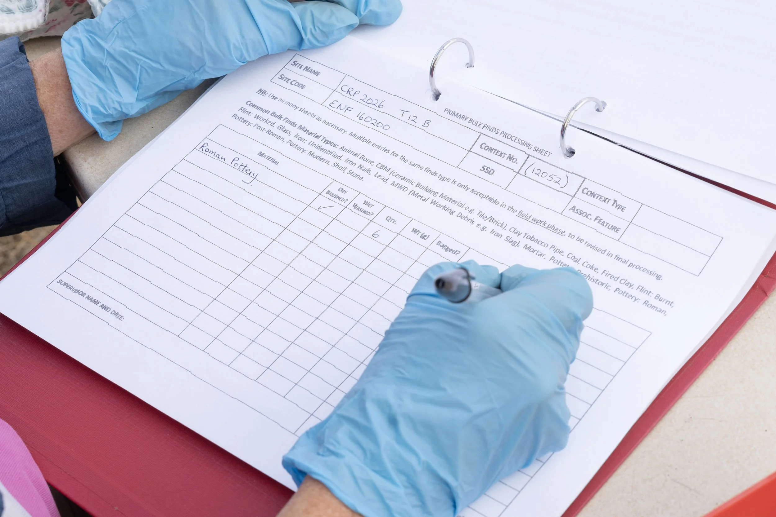 Close-up of an official document or processing sheet for archaeological artifacts, with handwritten notes, held by someone wearing blue gloves.