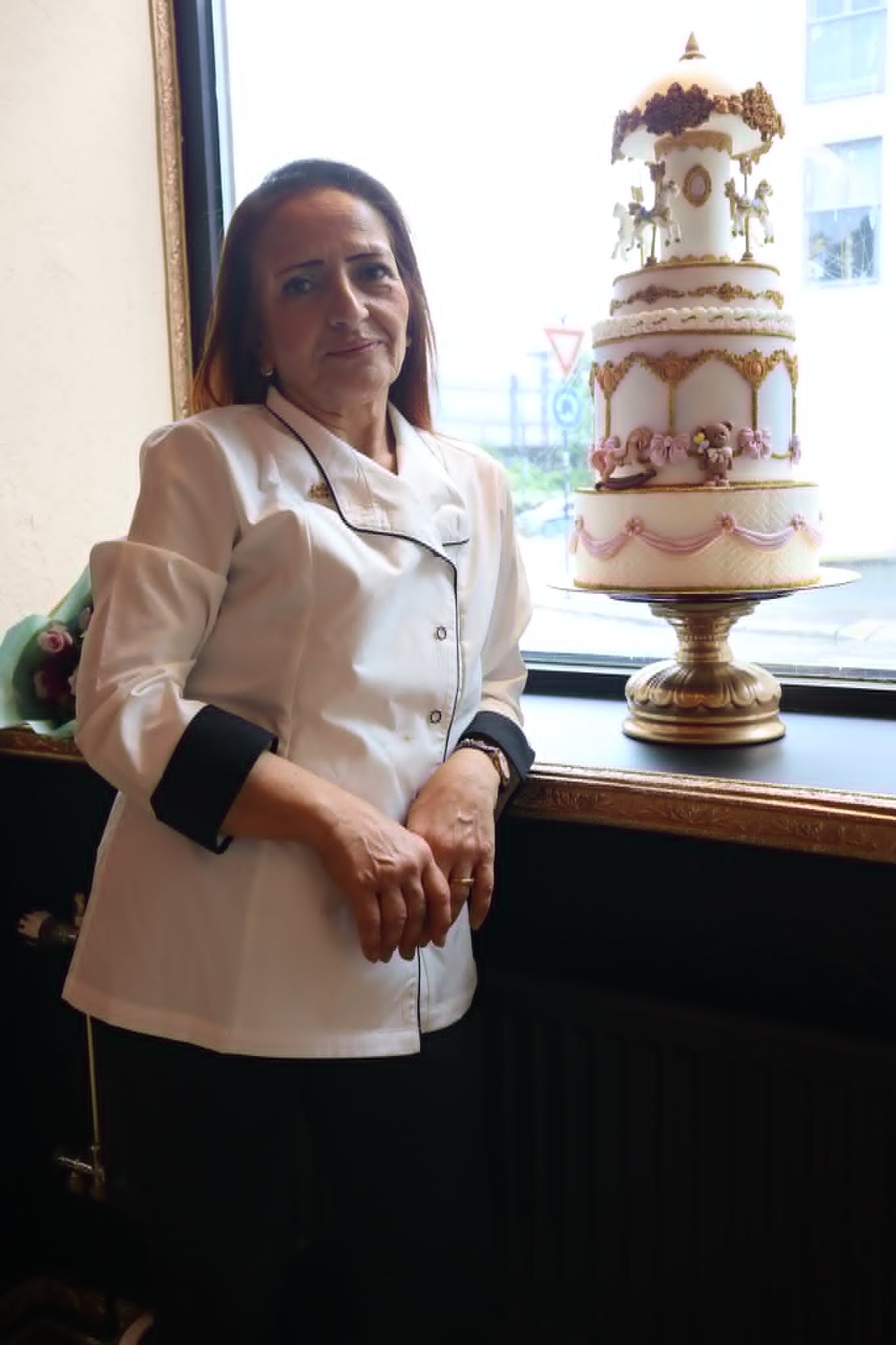 A woman in a white chef's coat standing next to a decorative multi-tiered cake with pink and gold details, displayed on a golden stand near a window.