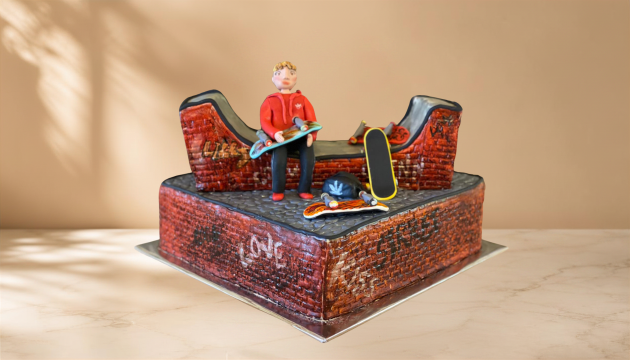 A skateboard-themed cake with a figurine of a boy sitting on a skatepark ramp, holding a skateboard, with additional skateboard accessories on top of the cake.