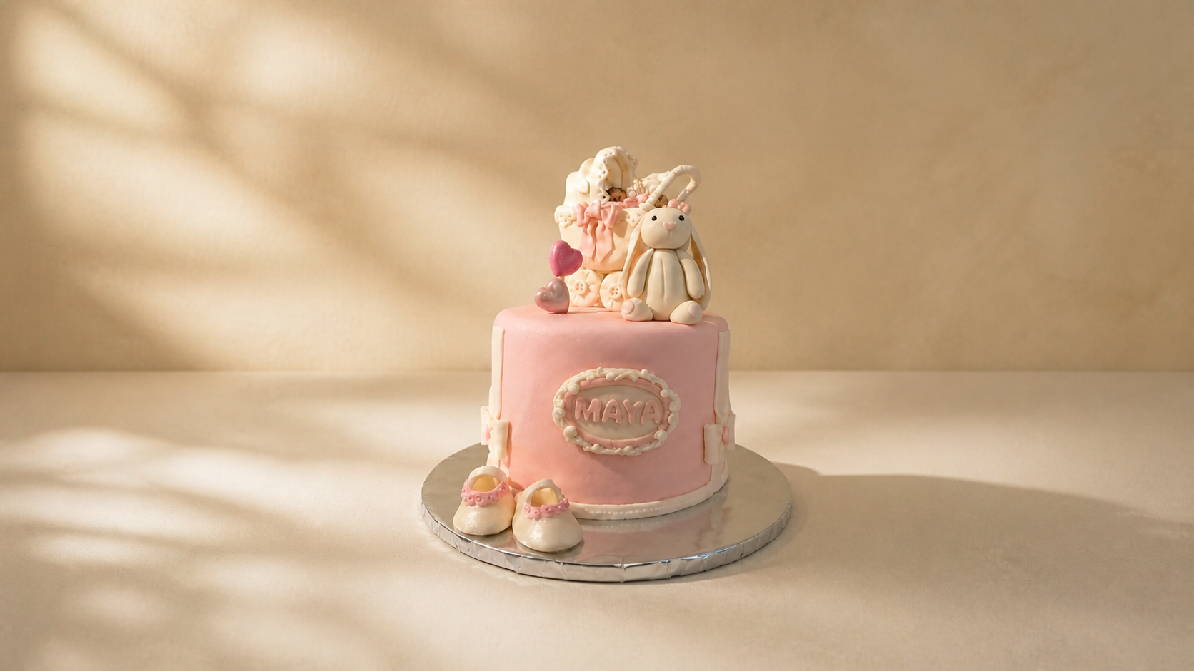 A pink birthday cake decorated with fondant baby shoes, a stuffed bunny, and heart-shaped decorations. The cake has a sign with the name 'Maya' on it and is set on a silver base with a beige background.