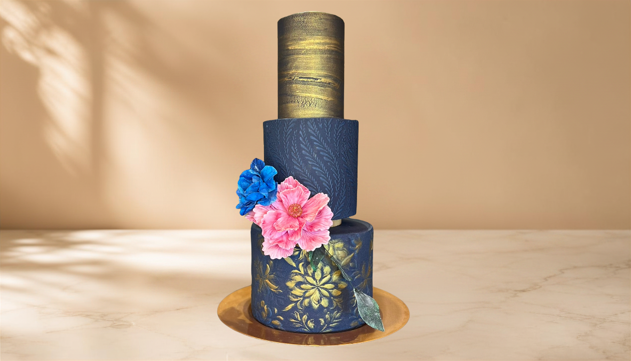 A decorative, three-tiered cake with gold and blue patterns, pink and blue flowers, and a gold top, placed on a gold-colored base on a light-colored surface.